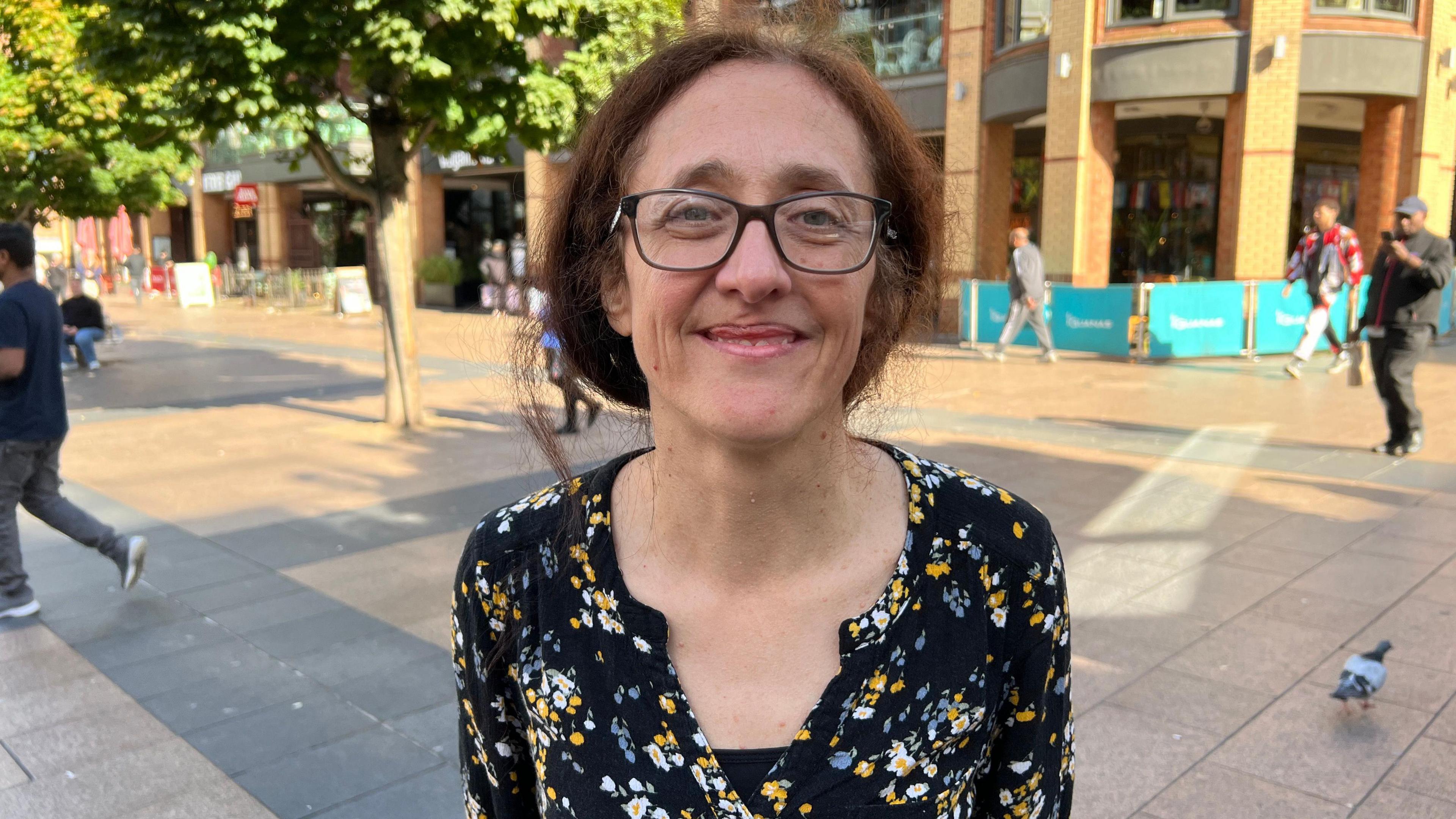 Joy Adams is pictured in Broadgate, in Coventry city centre, she is smiling at the camera and has glasses and is wearing a floral print blouse