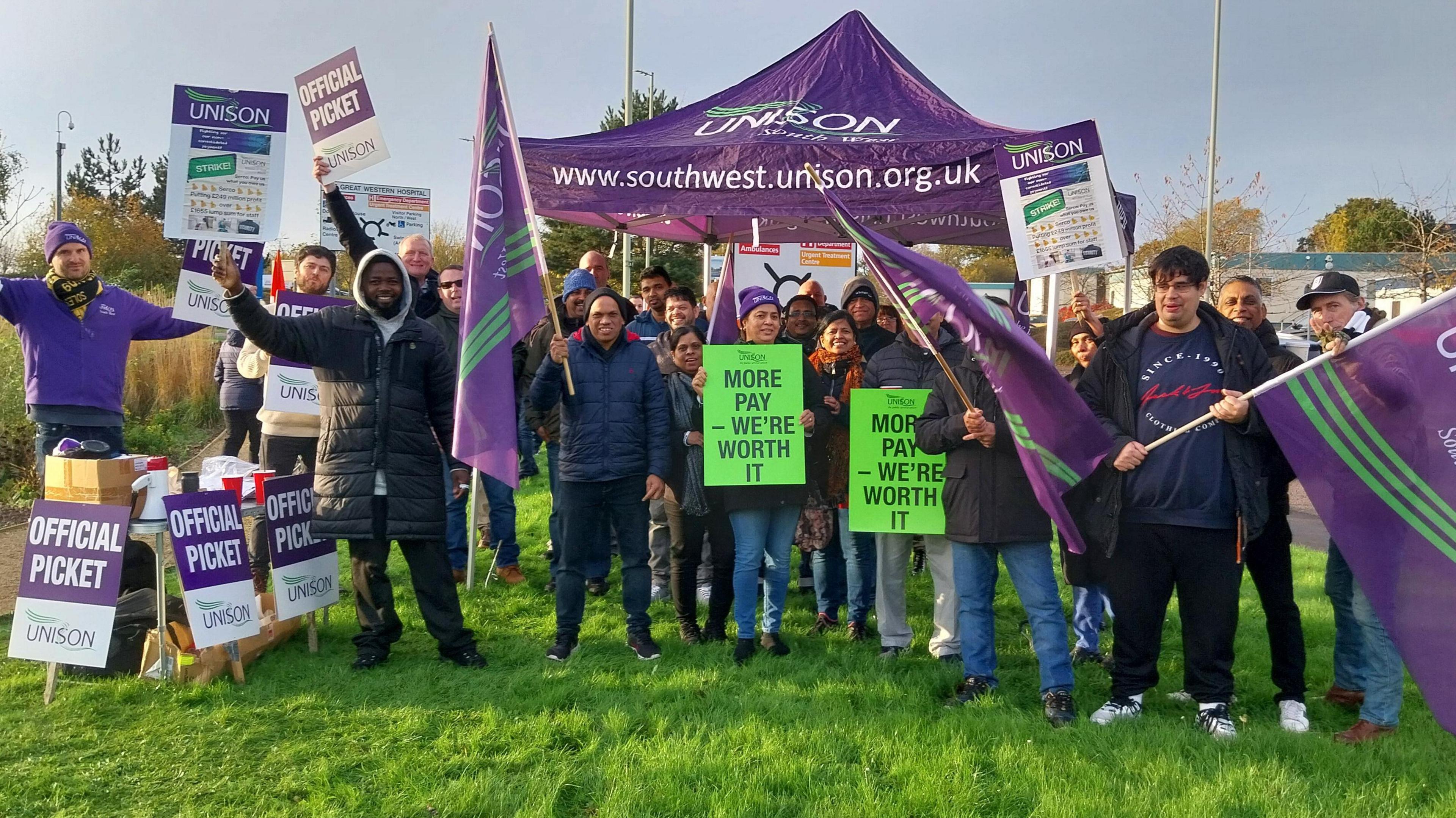 Strikers standing on grass with lots of purple unison signs and a gazebo. Purple flags also. Mostly wearing jackets and fleeces.