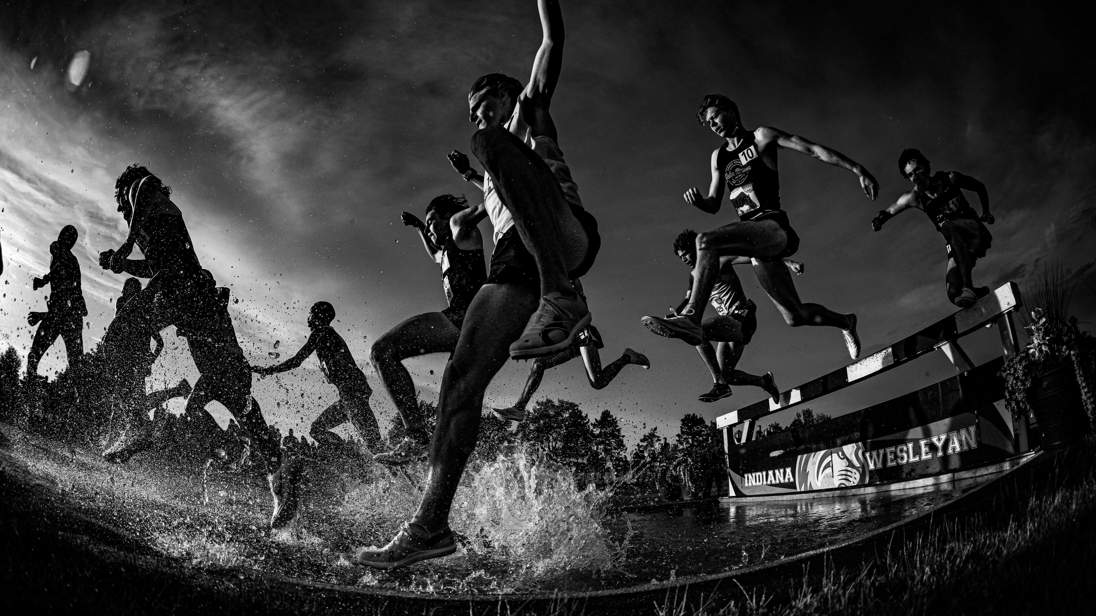 A group of male runners jump into the water pit during the steeplechase with a storm hovering over top of them