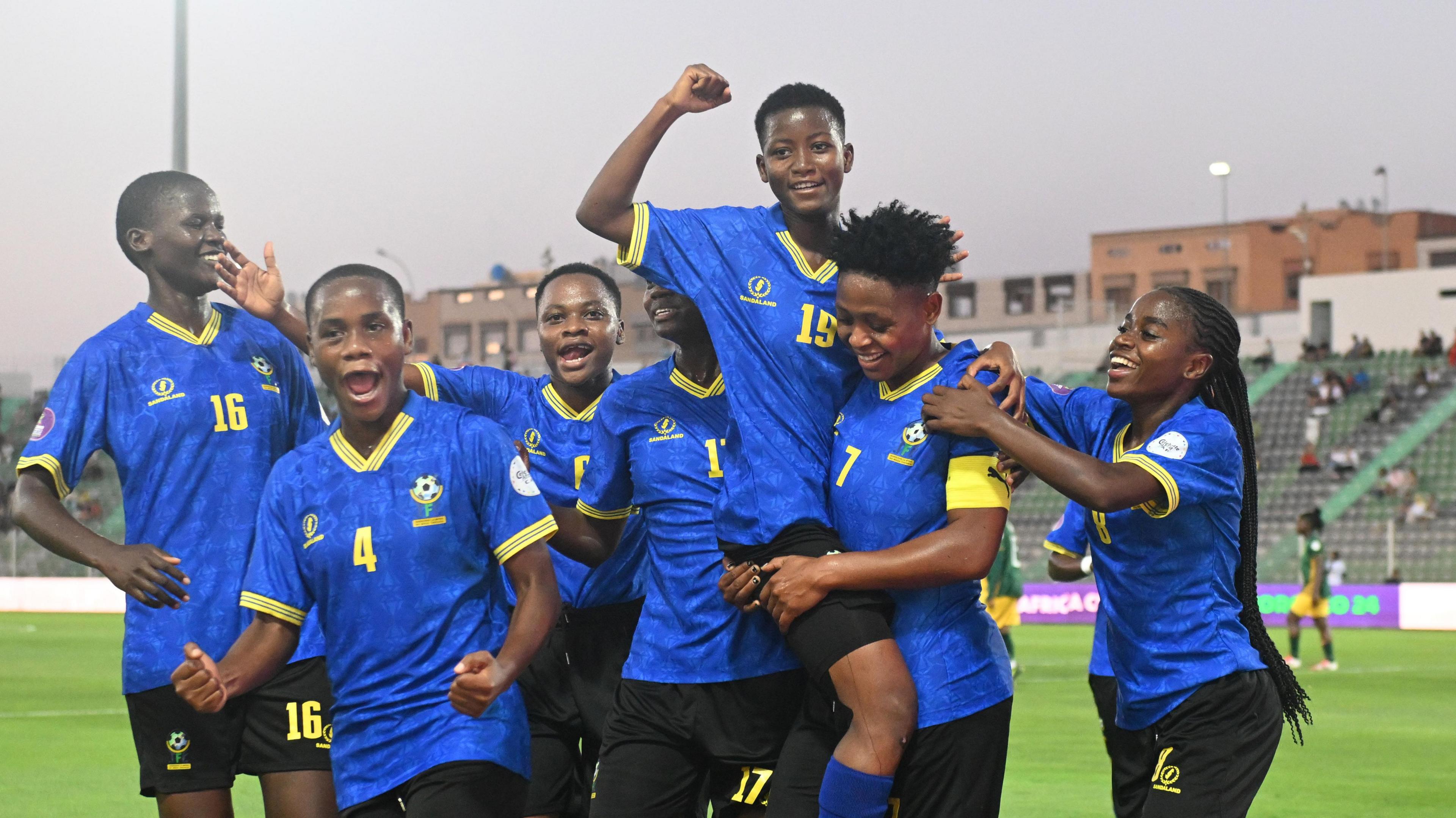 A group of seven Tanzania footballers wearing blue shirts with yellow trim and black shorts celebrate a goal, with one player raising a fist to the air while being carried by a team-mate