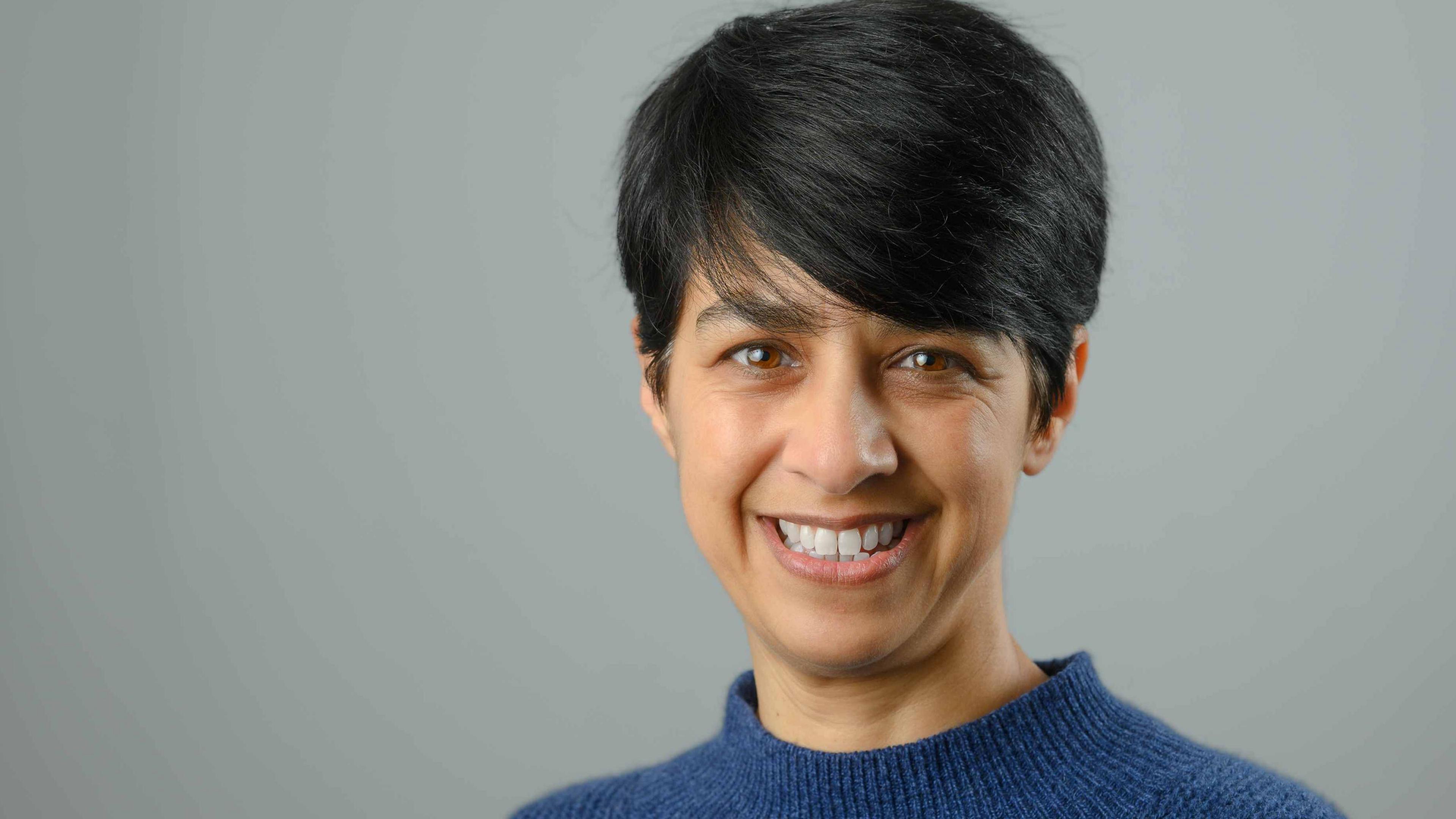 Professor Miranda Pallan smiling at the camera has dark hair and brown eyes. She is wearing a blue jumper.
