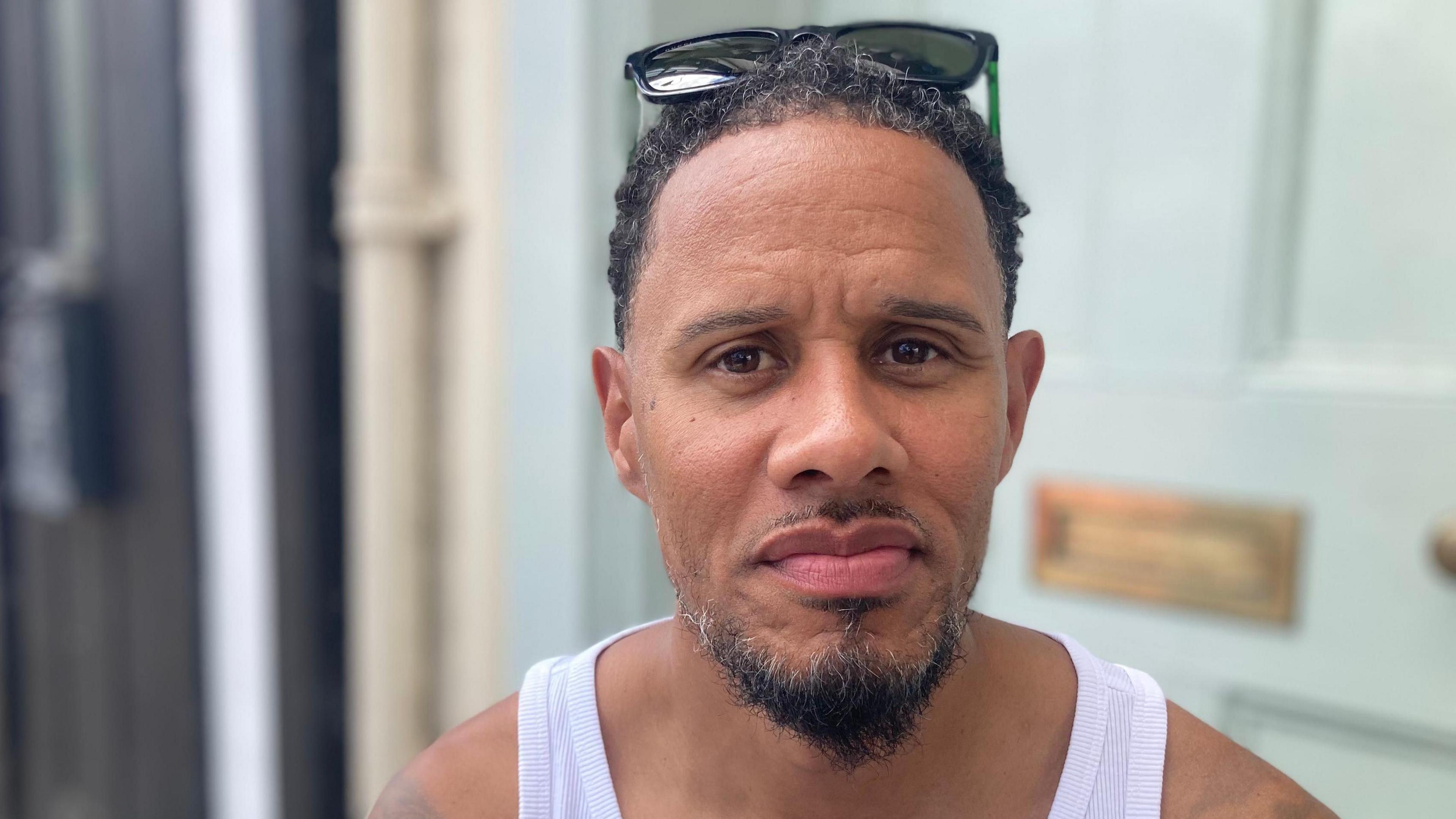 Anthony Burrell is looking towards the camera. He has short black and grey hair, and has sunglasses on his head. He is wearing a white tank top and is standing in front of a very pale blue door.