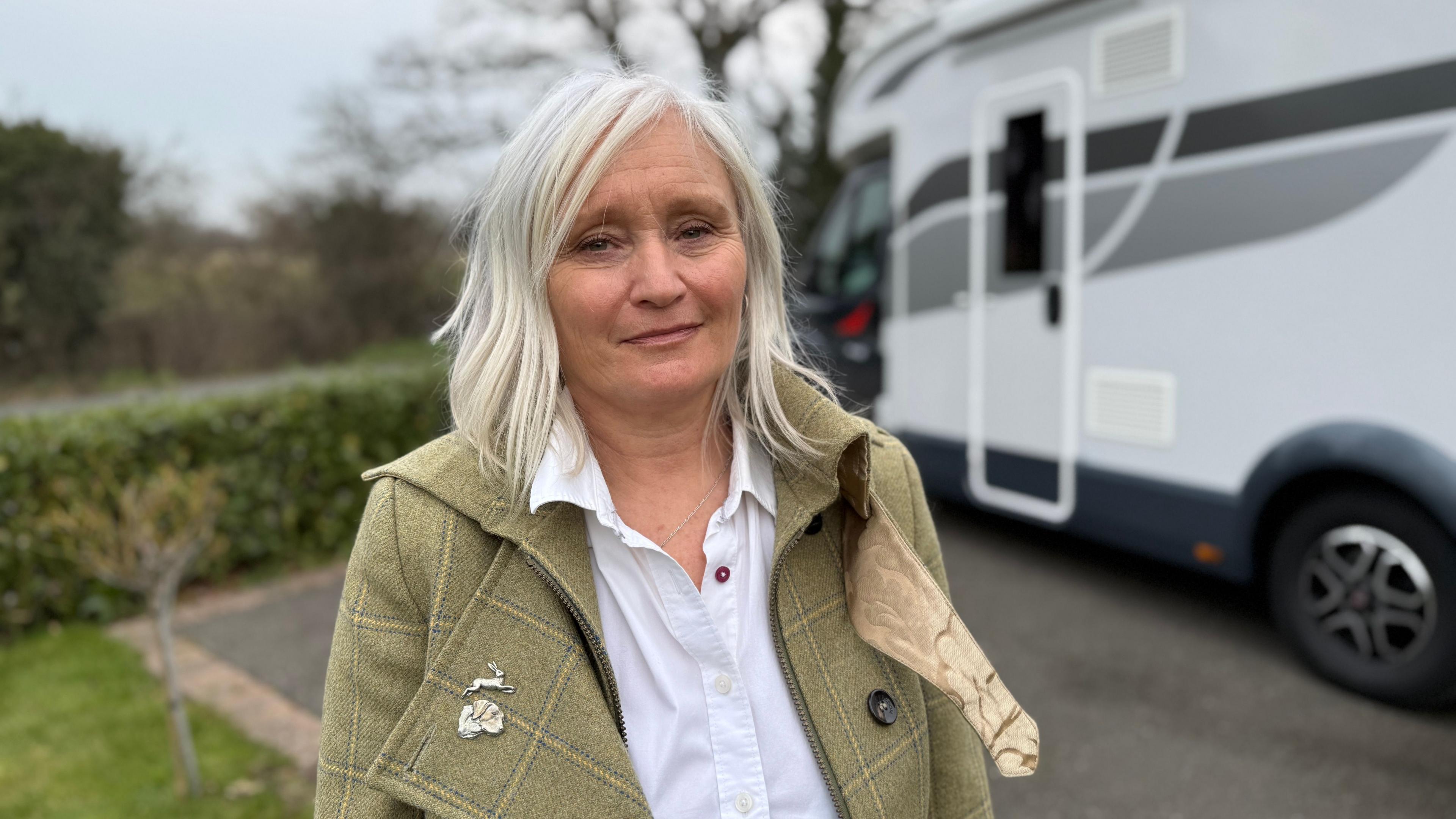 Joanne Fenton with long blond hair and stone-coloured jacket with white shirt underneath. She is looking at the camera. She is beside a white motorhome with grey and black detailing.