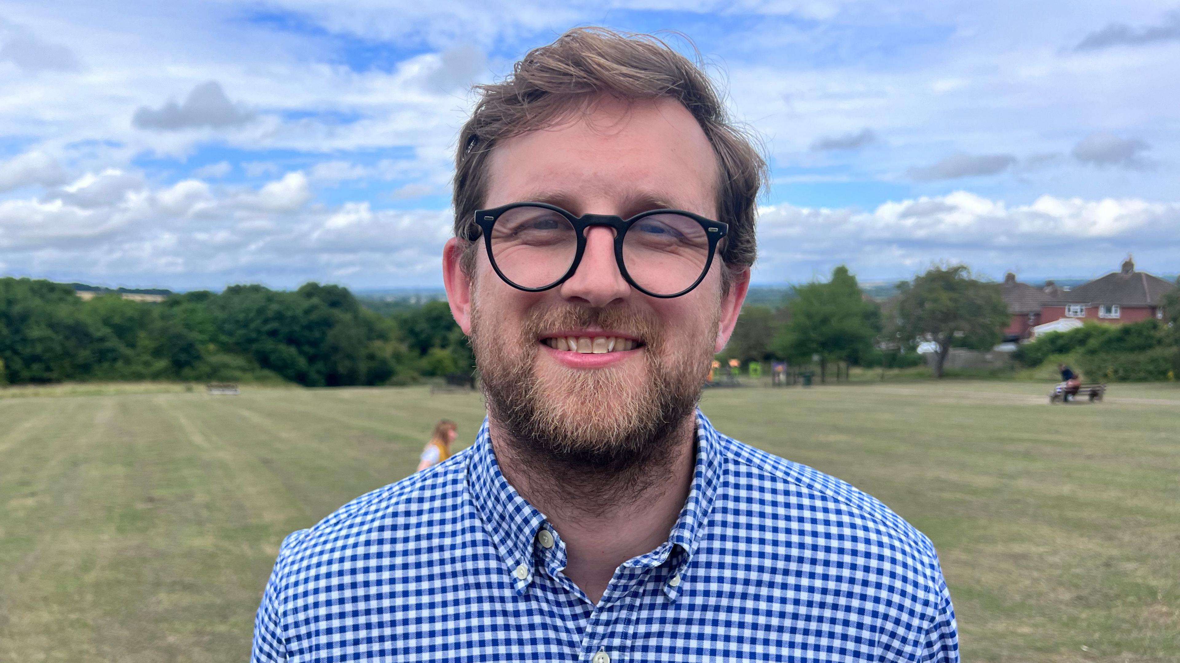 Freddie van Mierlo has short brown hair, a brown beard, and is wearing glasses and a blue and white plaid shirt. Behind him out of focus is a green field and trees.