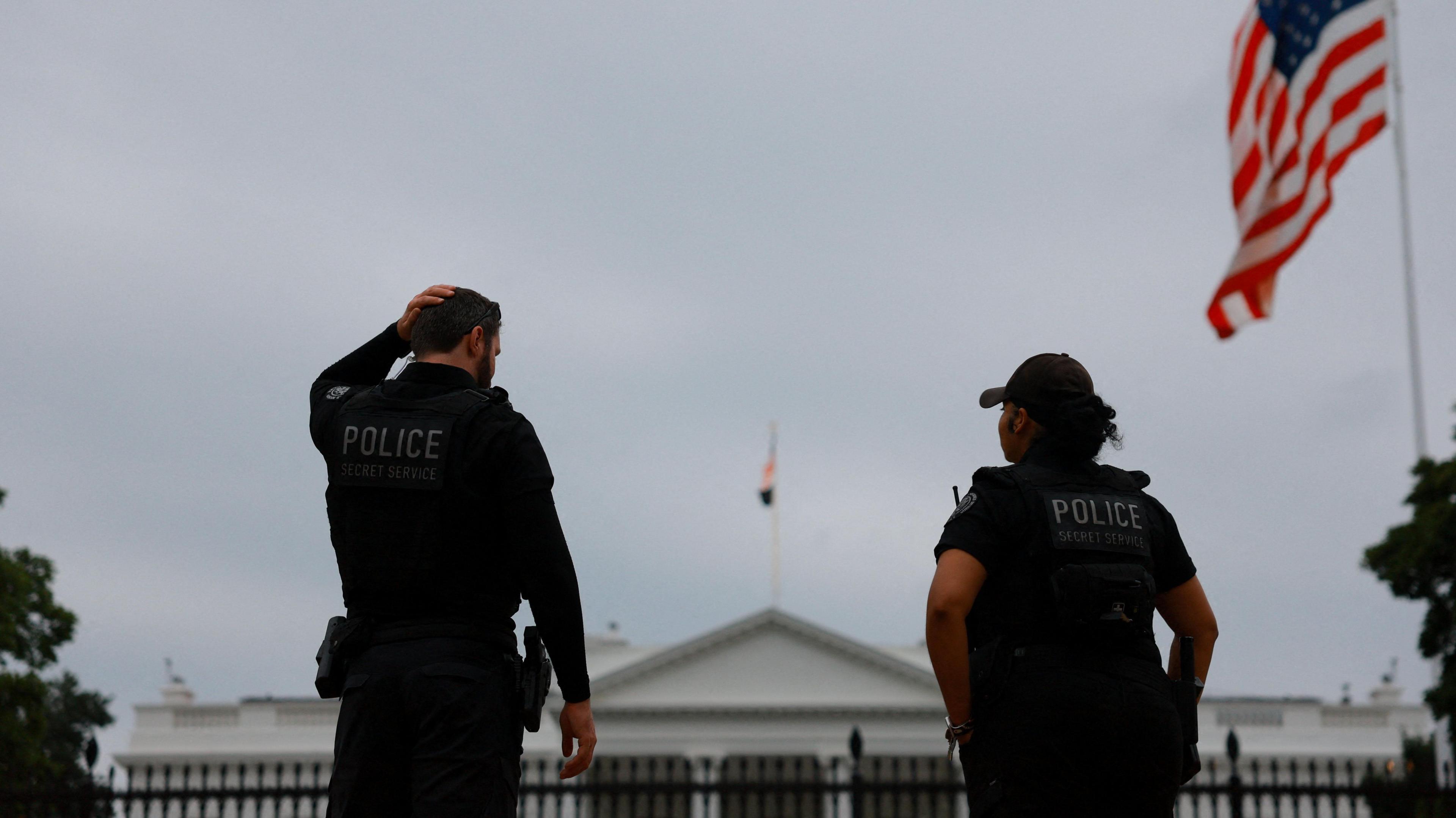 Two police officers stand facing the White House with their backs to the camera. A US flag flies on the right.