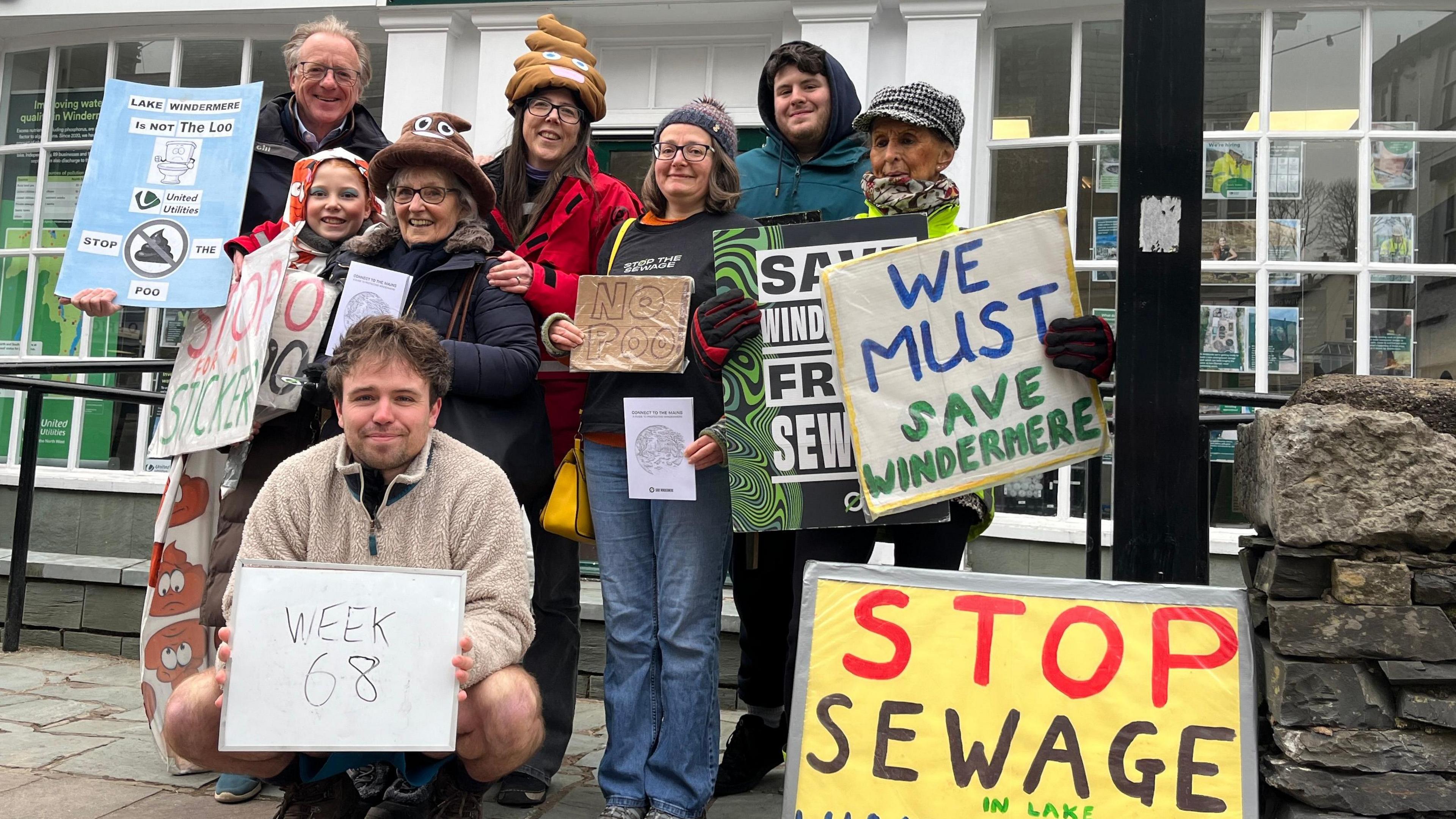 A group of anti-sewage campaigners pose for a photo. They are holding signs saying things such as "Stop Sewage" and "We must Save Windermere".