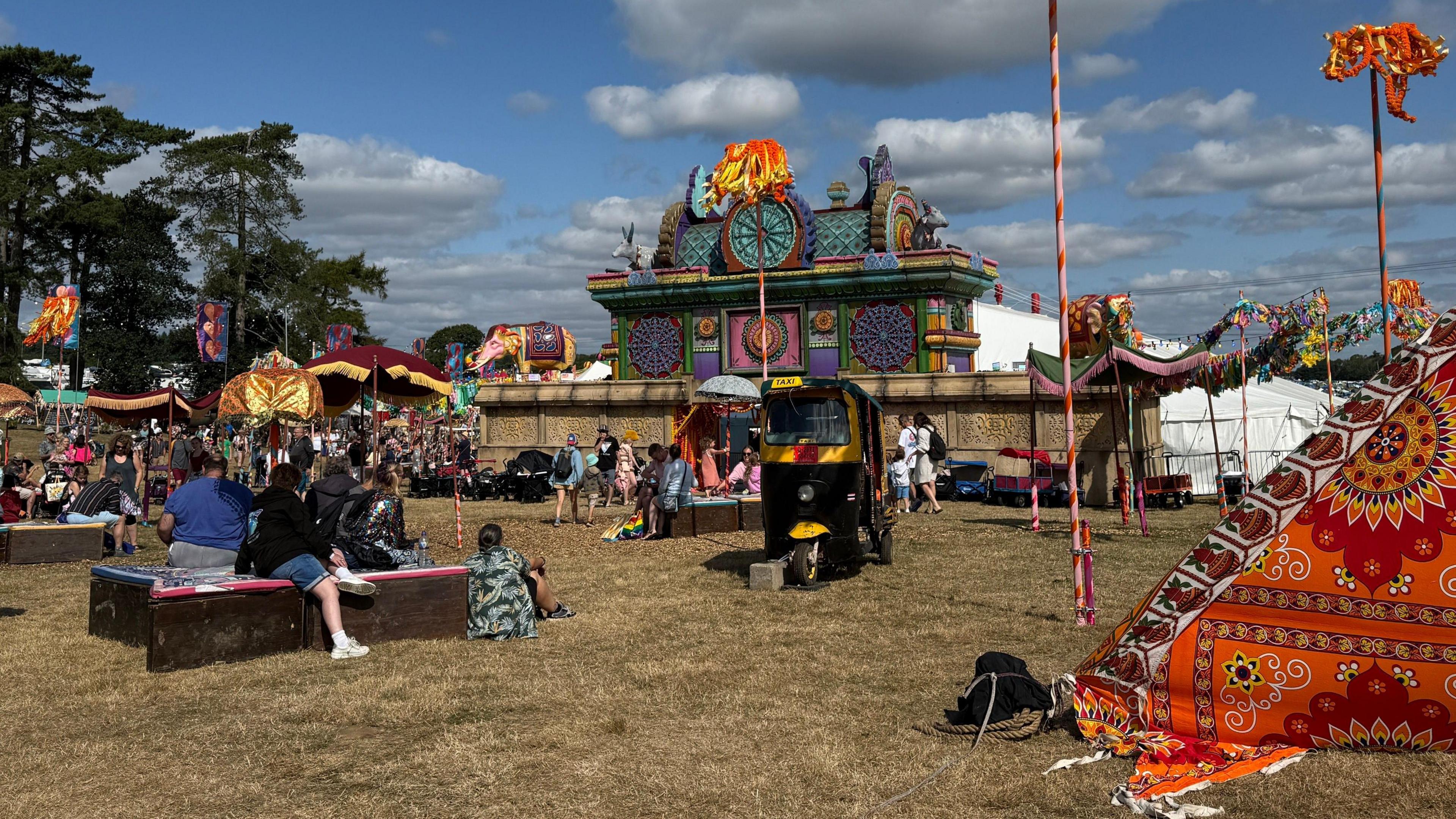 A large colourful building is surrounded by colourful tents and flags. Festival-goers sit on wooden blocks around it.