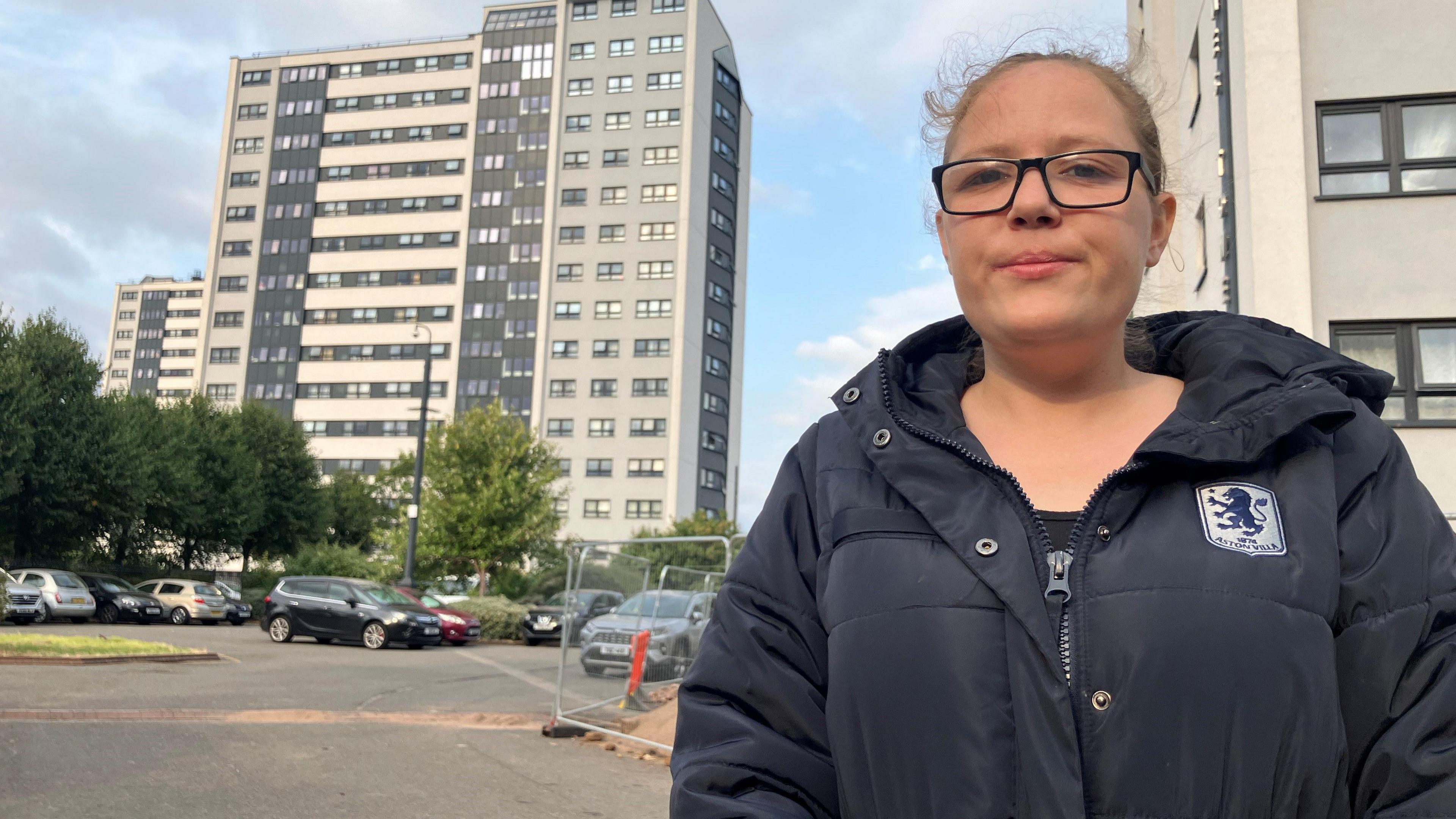 Woman looking down at camera in front of tower blocks. She is wearing a black puffa coat and black framed glasses, with tied-back fair hair.