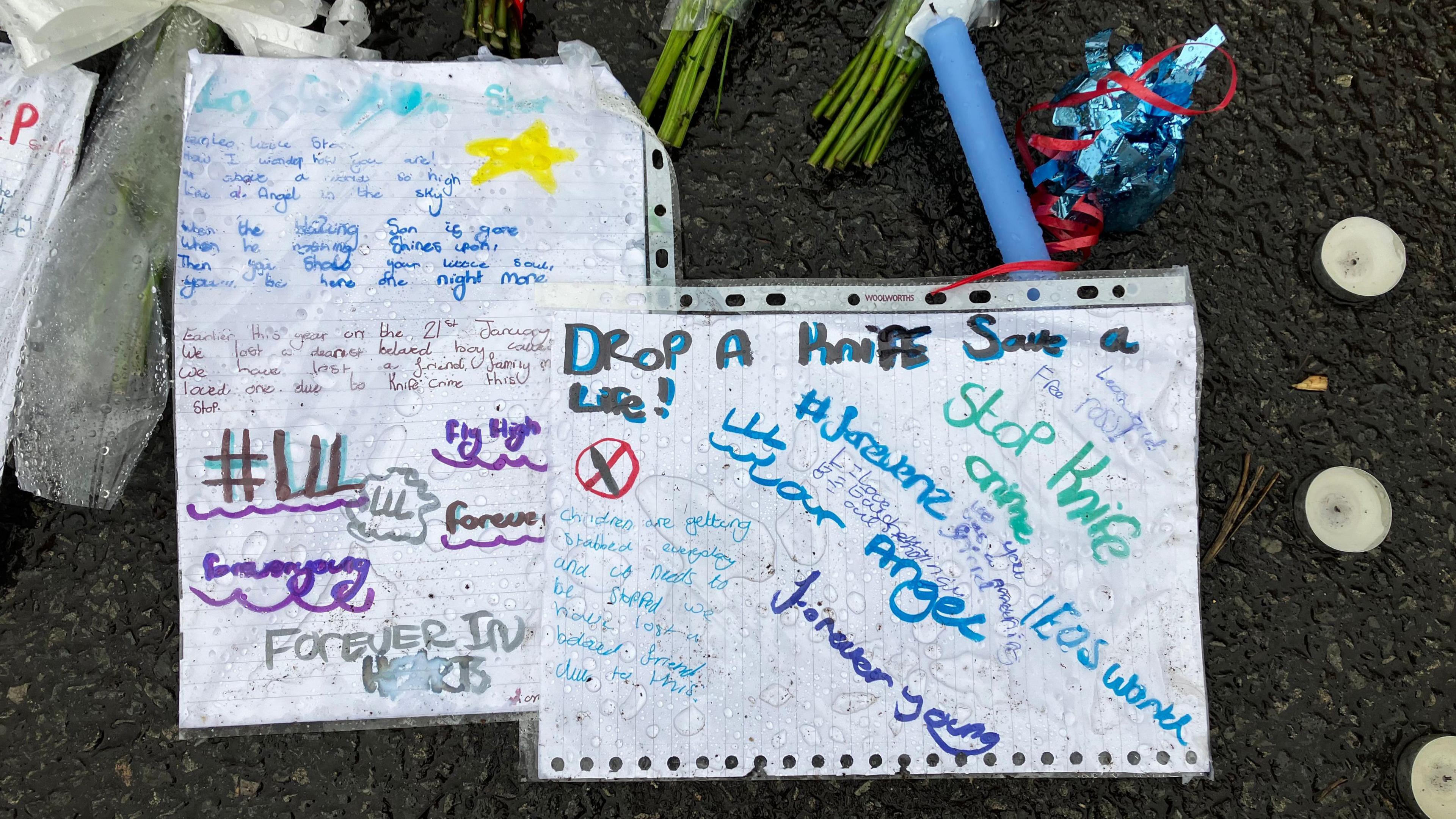 Pieces of white paper, inside plastic ring binder sheets, carry messages such as 'drop a knife, save a life' and are placed alongside flowers and candles.