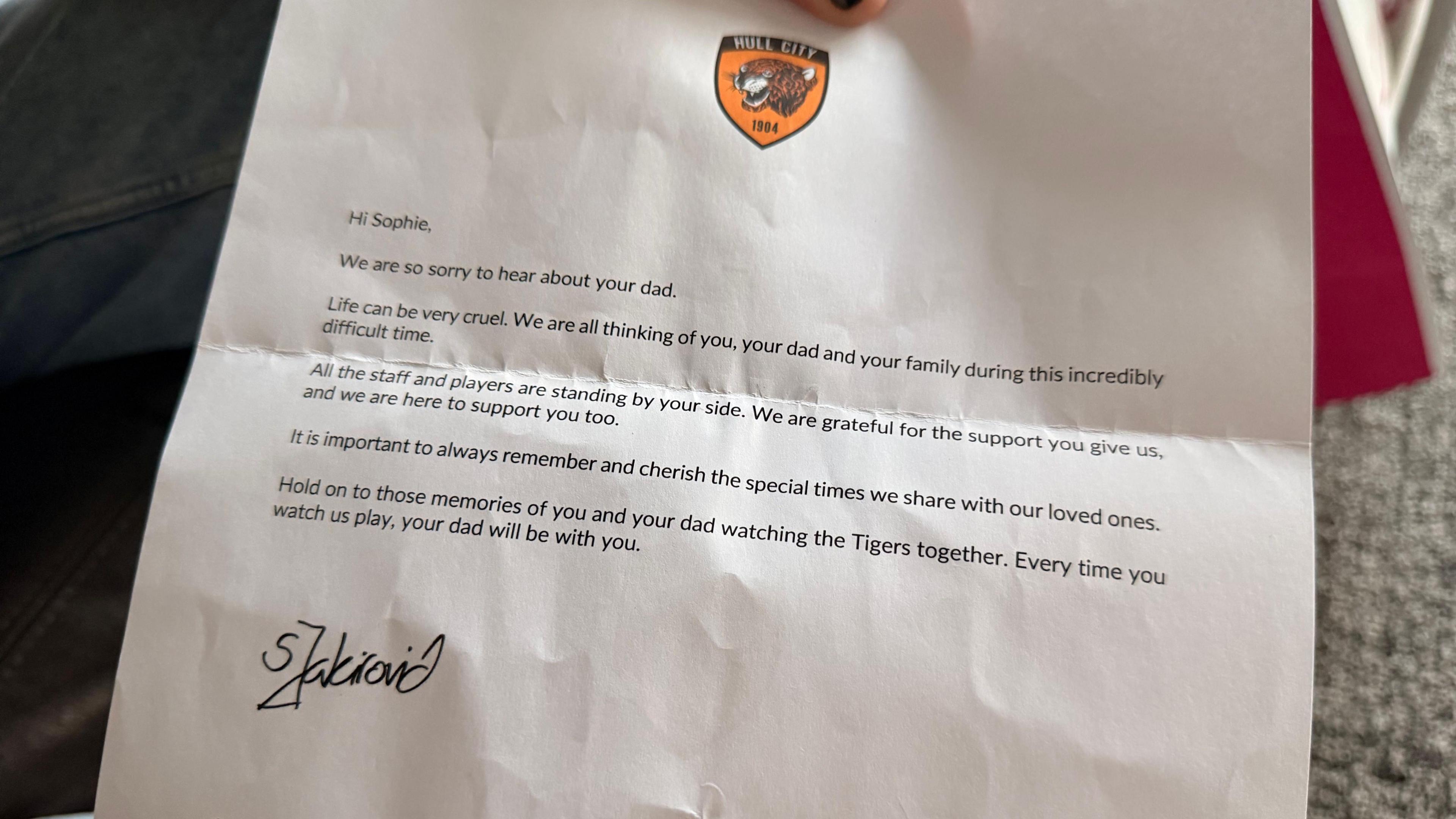 A letter on white paper. It has a Hull City FC badge at the top. It reads: "To Sophie, We are so very sorry to hear about your dad. Life can be so very cruel. We are all thinking of you, your dad and your family during this incredibly difficult time. All the staff and players are standing by your side. We are grateful for the support you give us, and we are here to support you too. It is important to always remember and cherish the special times we share with our loved ones. Hold on to those memories of you and your dad watching the Tigers together. Every time you watch us play, your dad will be with you."