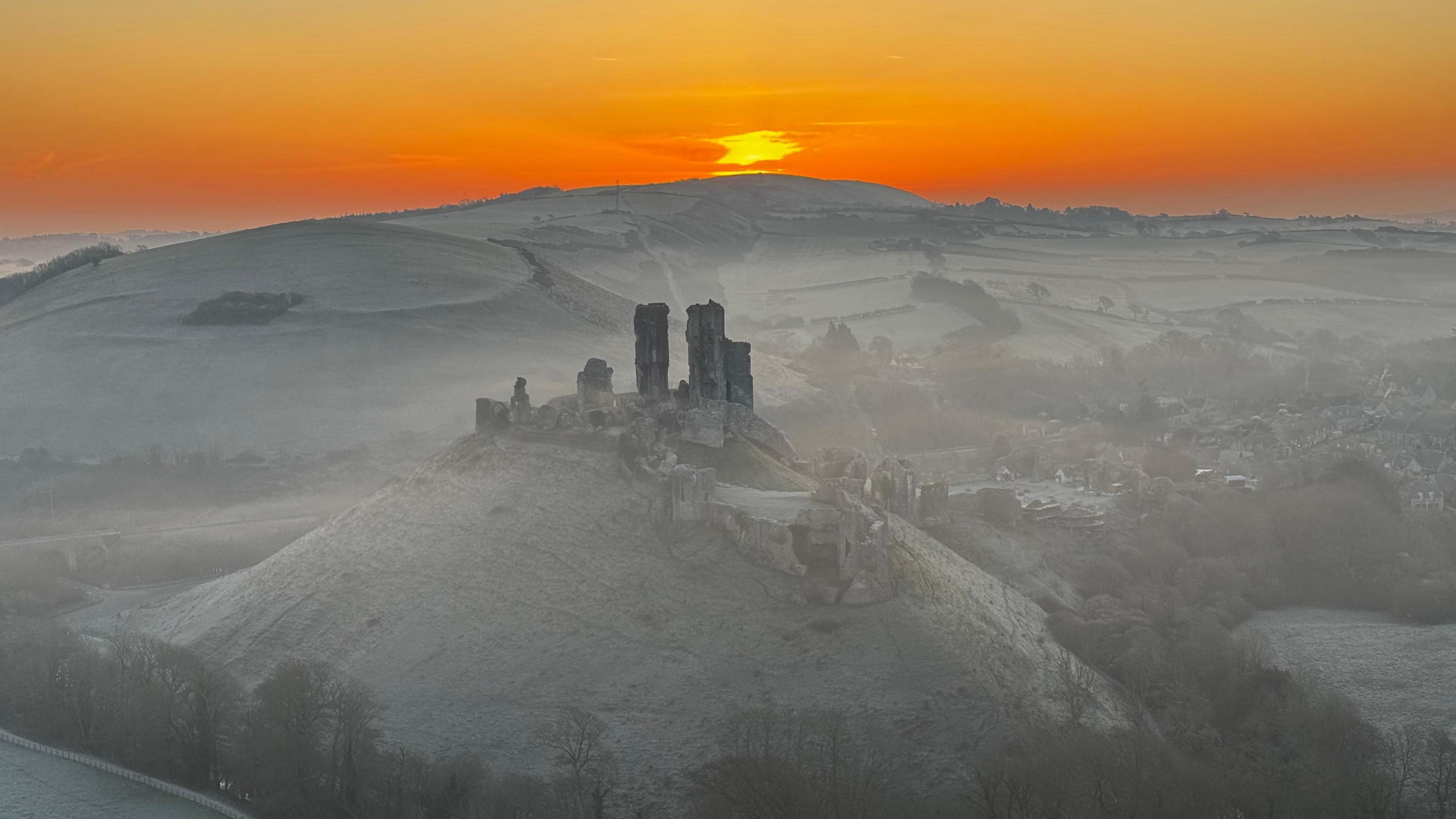 The ruins of Corfe Castle stand on a hill in the Dorset countryside. The surrounding fields are covered in a white frost. The sky is glowing orange as the sun appears over the horizon.