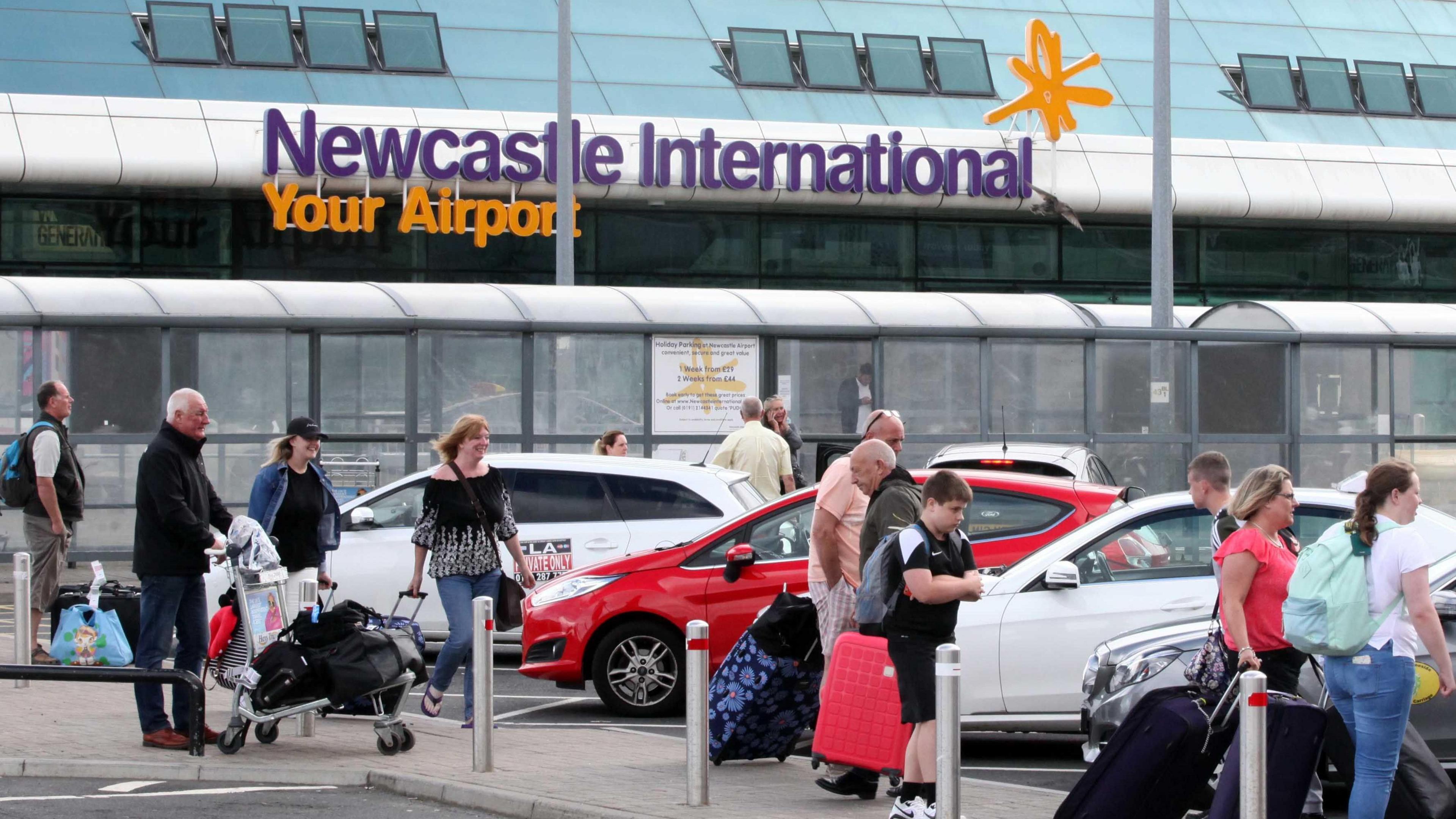 Newcastle International Airport with purple and yellow sign. Passengers walking away from the building with luggage.