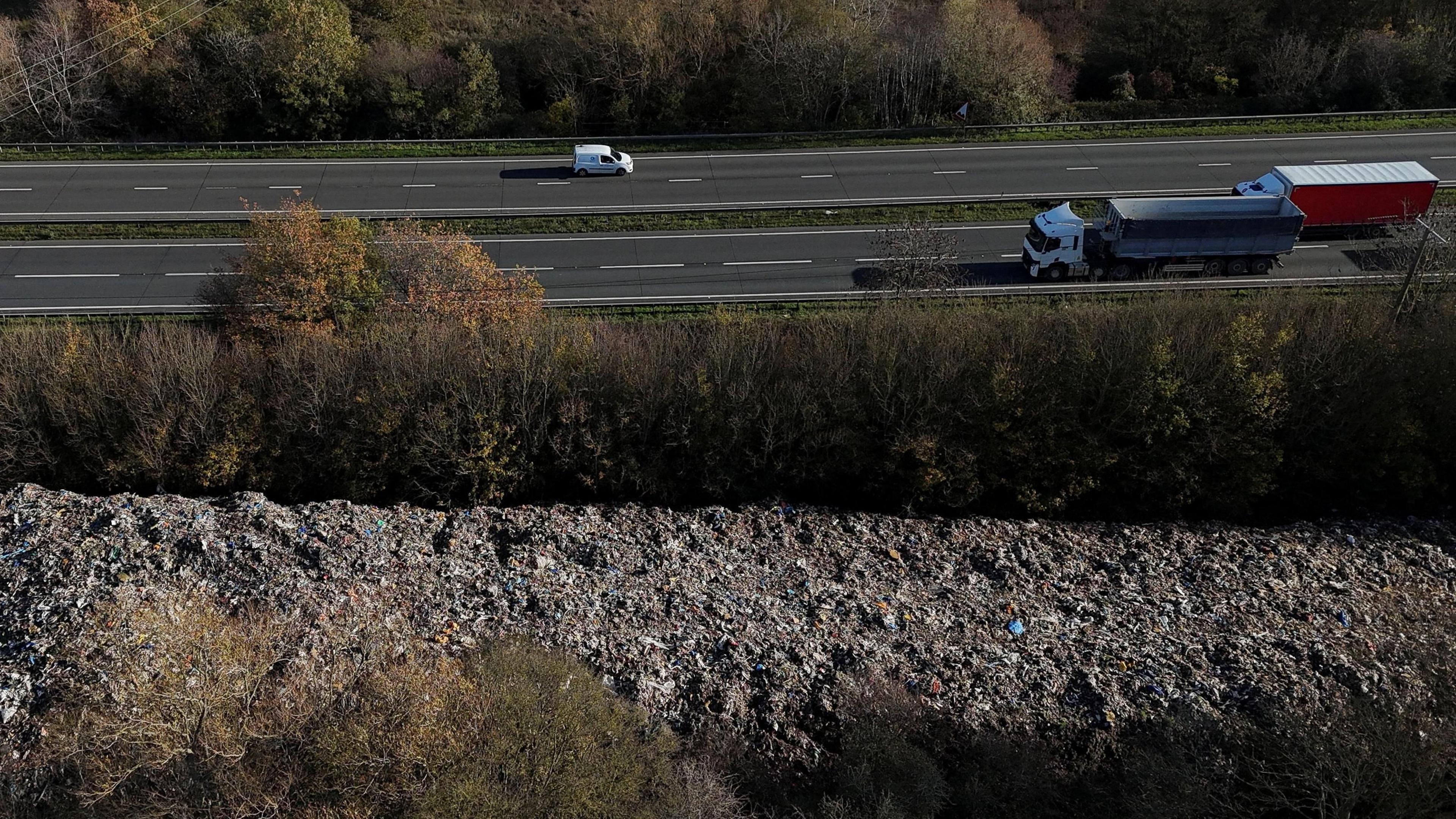 A dual carriageway running across the top of the image with a parallel pile of rubbish at the bottom of the image, with trees in between.