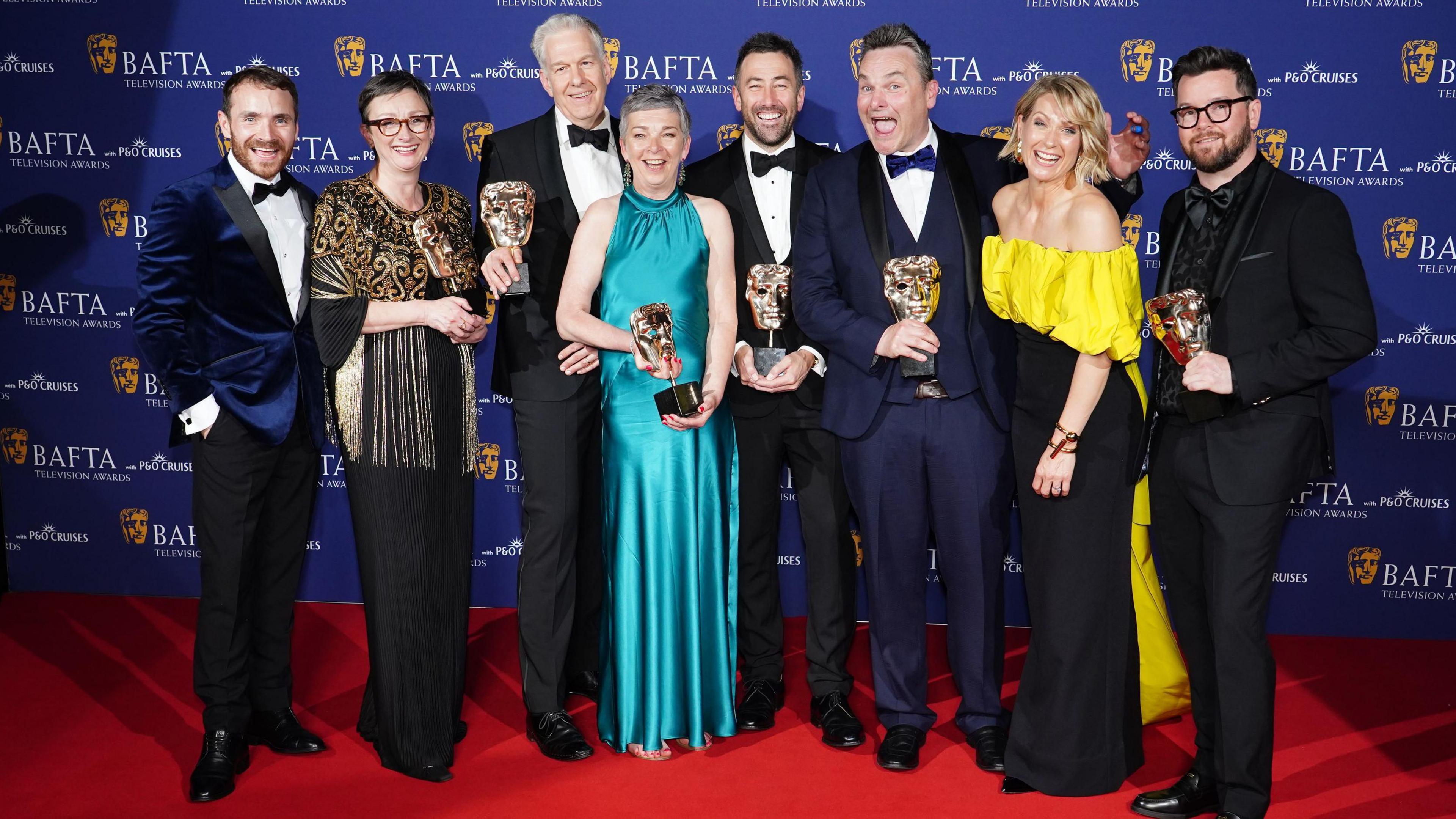 Martin McCann, Louise Gallagher, Stephen Wright, Amanda Black, Adam Patterson, Declan Lawn, Sian Brooke and Jack Casey in the press room after winning the Drama Series Award for 'Blue Lights' at the 2025 BAFTA TV Awards in London. They are stood on a red carpet holding their awards and smiling at the camera.
