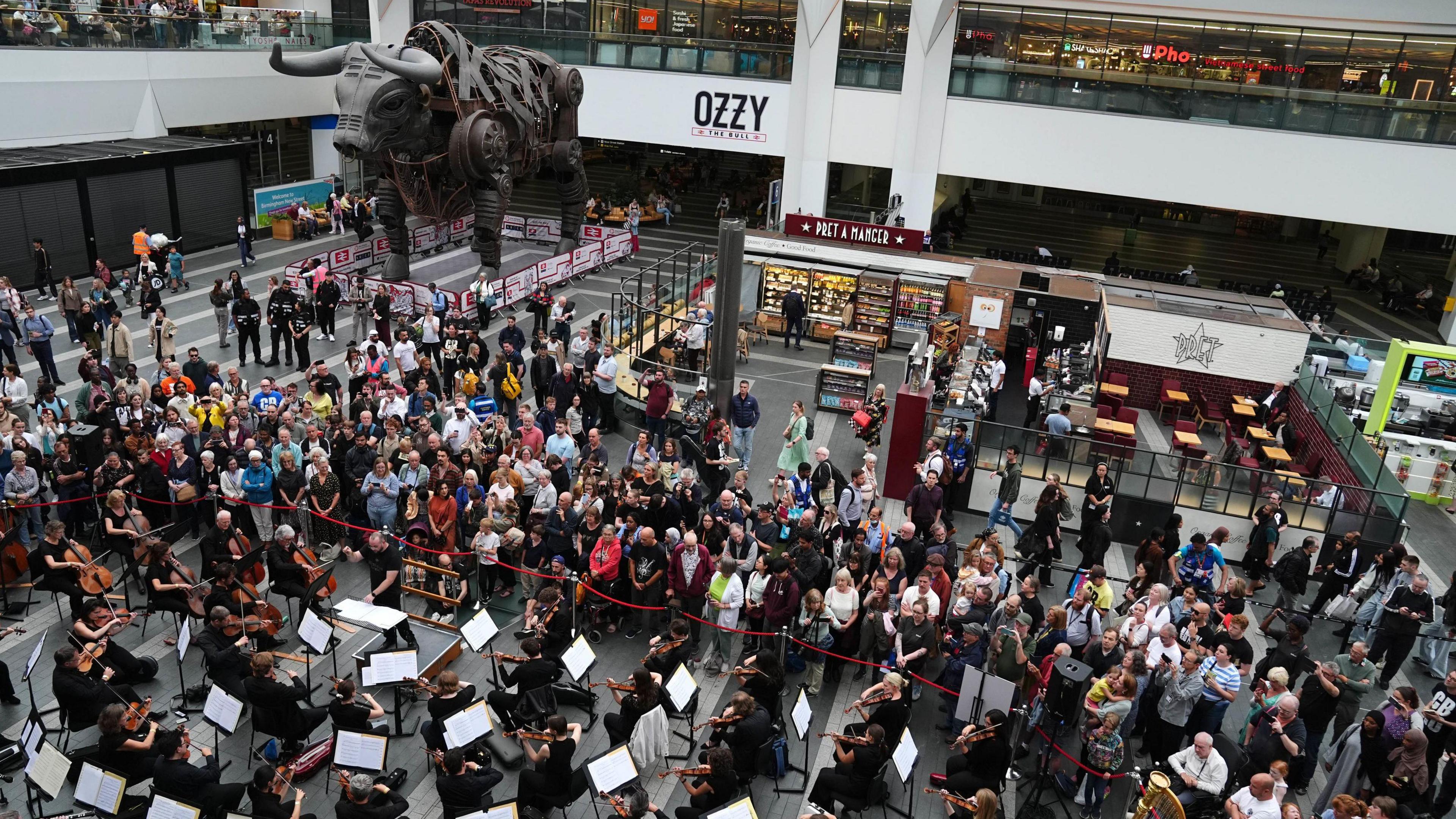 A large orchestra performing in front of a large crowd next to a large bull statue at a train station.