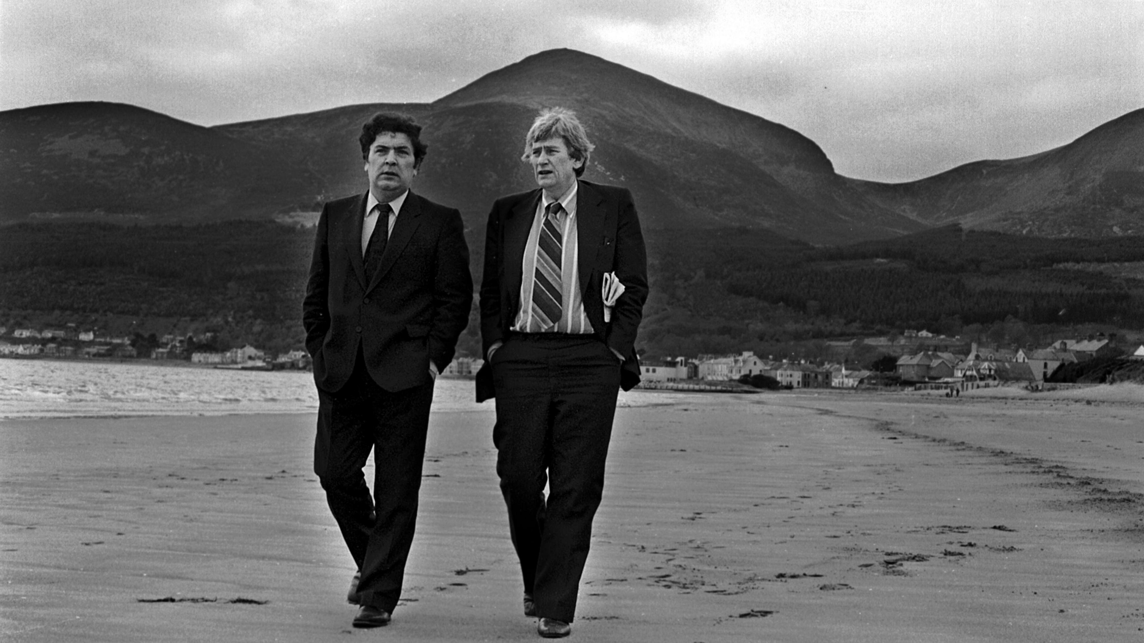 A black and white image of two men walking across the beach. One has dark short hair and the other lighter hair. Both are wearing a suit with tie and shirt. The Mourne mountains are pictured in the background, and the waves are crashing on the shore behind them.