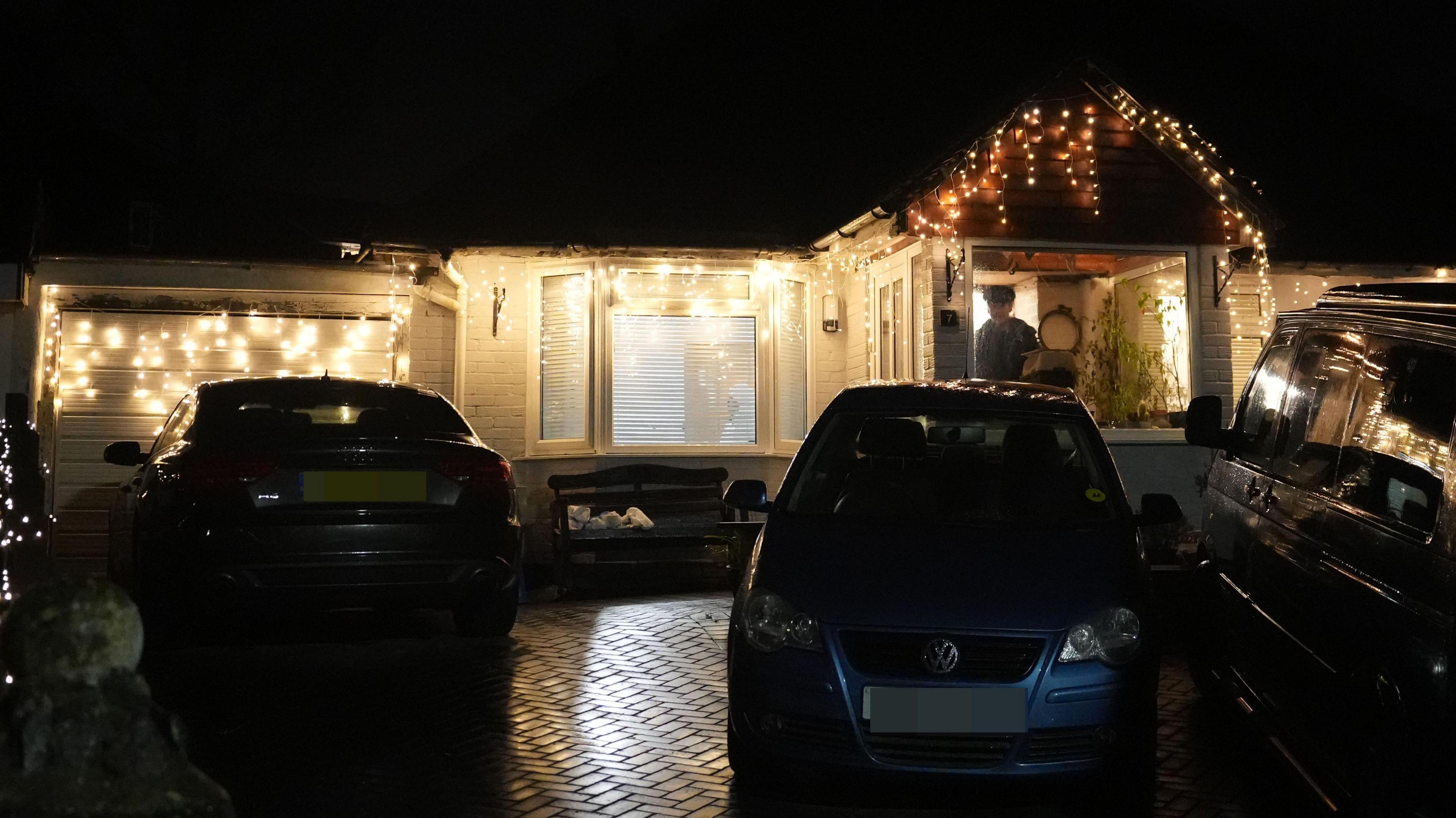 Two blue cars on a drive outside a white house with Christmas lights on