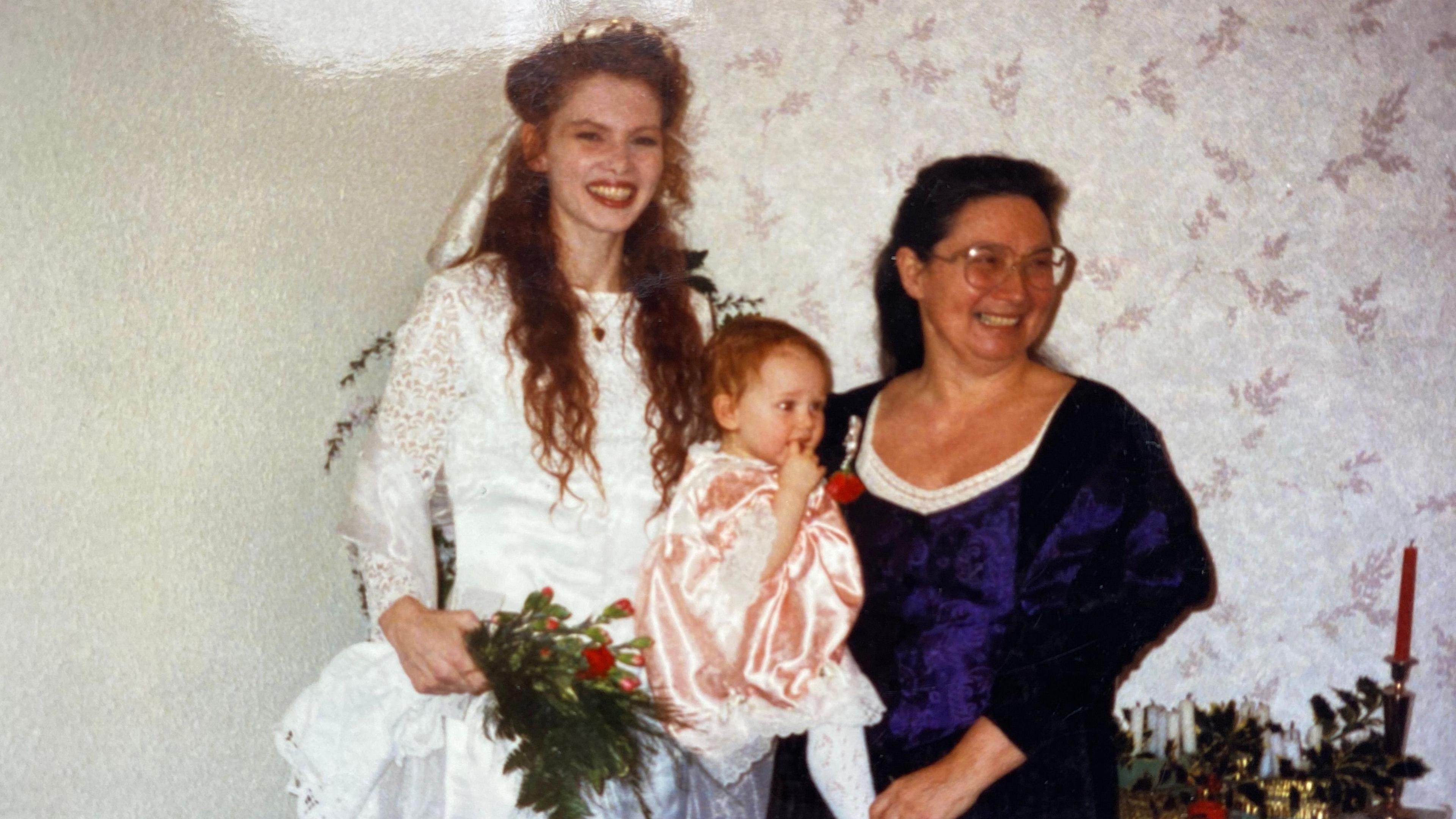 Alicia with her daughter Janie and her granddaughter Josie on Janie's wedding day. Alicia wears a dark blue dress, Janie wears a long-sleeved lacy bridal gown and Josie, a young baby, wears a pink dress.
