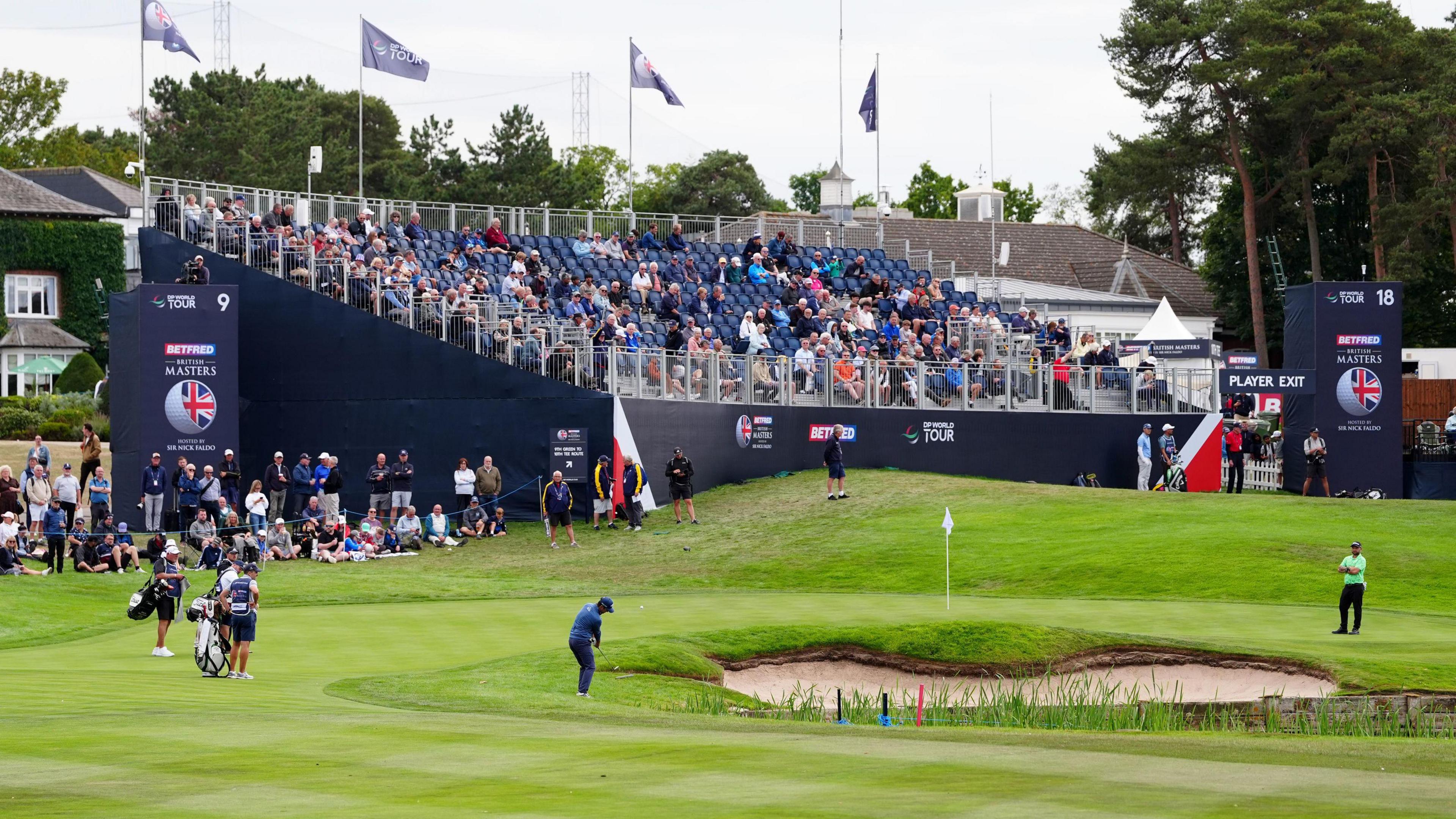 Golfers are scene on the green of The Belfrey. There is a bunker in the green, and a blue stand full of spectators in the background.