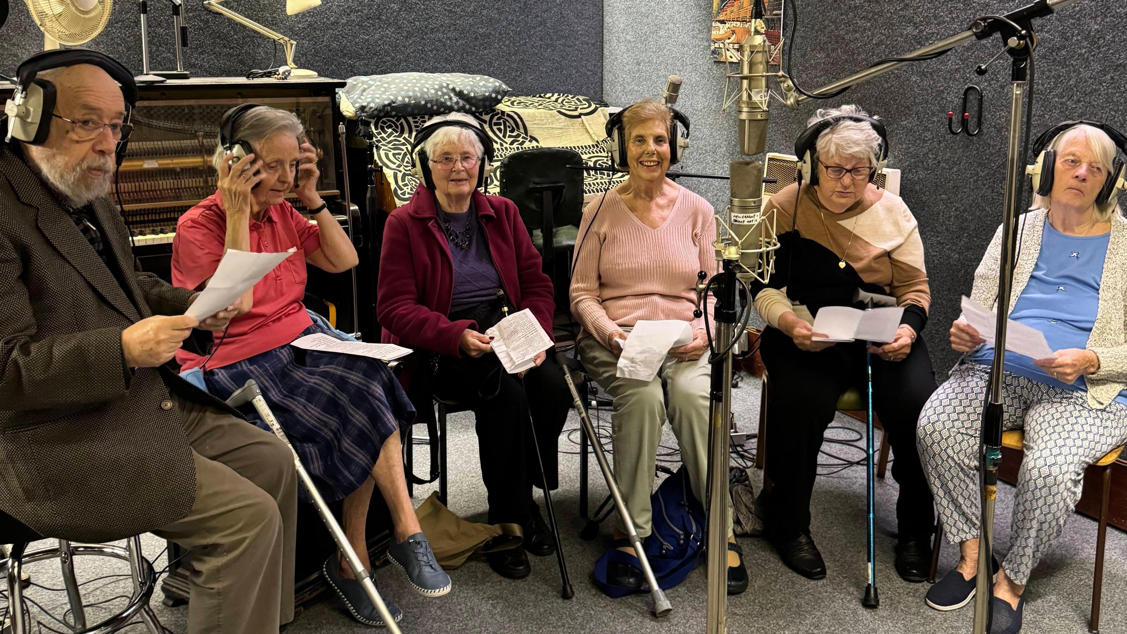 A man and five women, all residents at a care home, sit around microphones in a recording studio. Behind them is a brown upright piano and grey soundproofed walls.