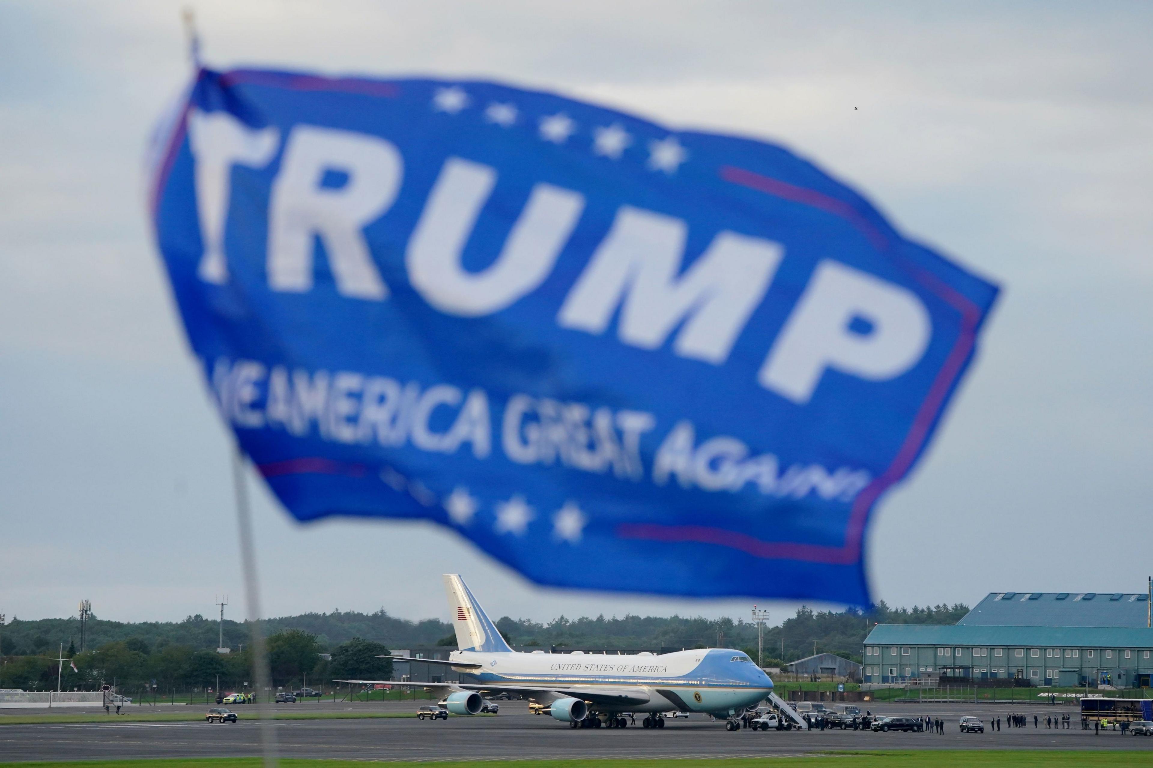 Air Force One is on the tarmac in the distance. In the foreground is a blue flag with the words Trump and Make America Great Again.