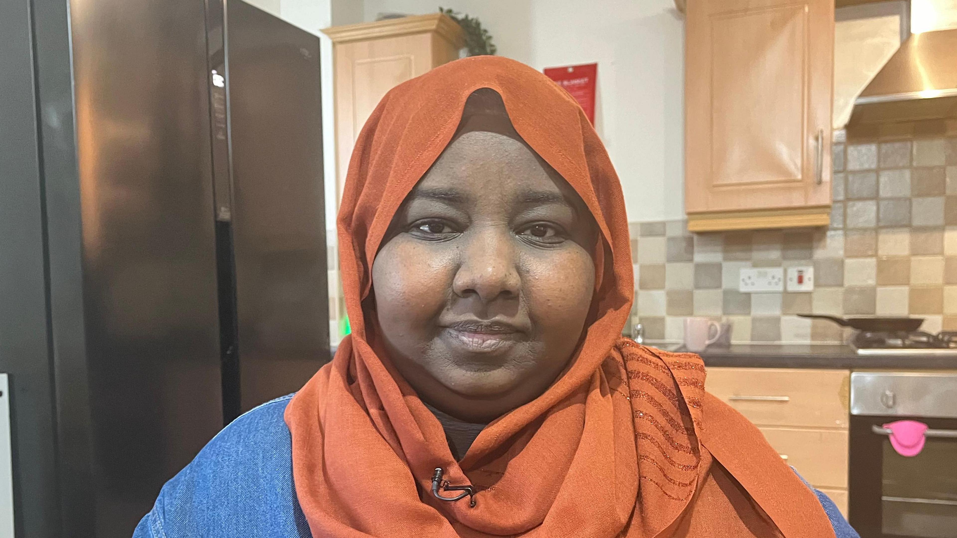 Suha, wearing an orange head scarf and a denim shirt, with a kitchen in the background