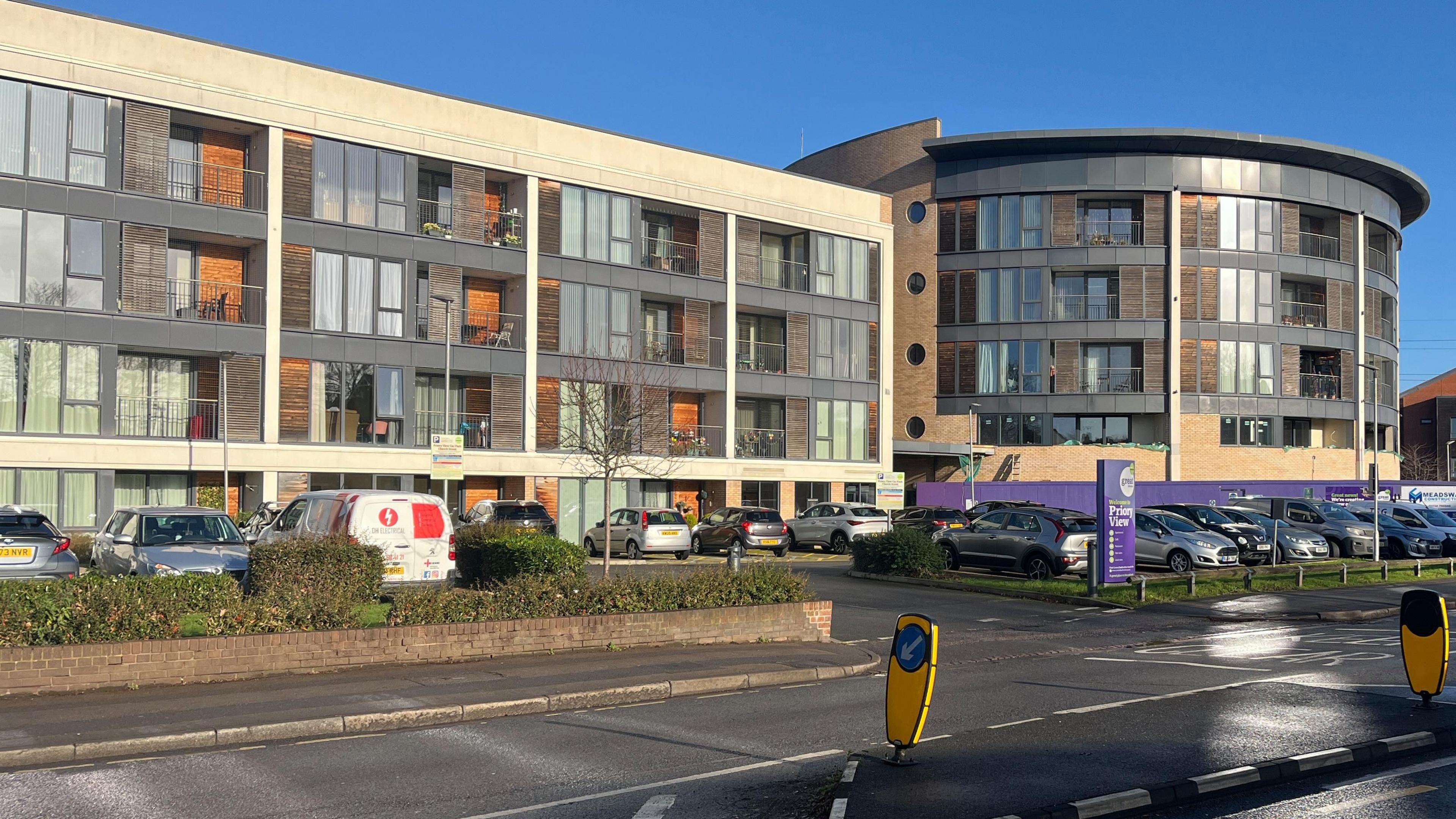 A modern multi-story building complex with a mix of rectangular and curved architectural designs. The left section of the building has three floors with large glass windows and wooden panel accents, while the right section is a rounded structure with similar glass and wood detailing. In front of the building, there is a parking area filled with several cars and a white van. A purple sign and a purple wall or barrier are visible near the entrance area.