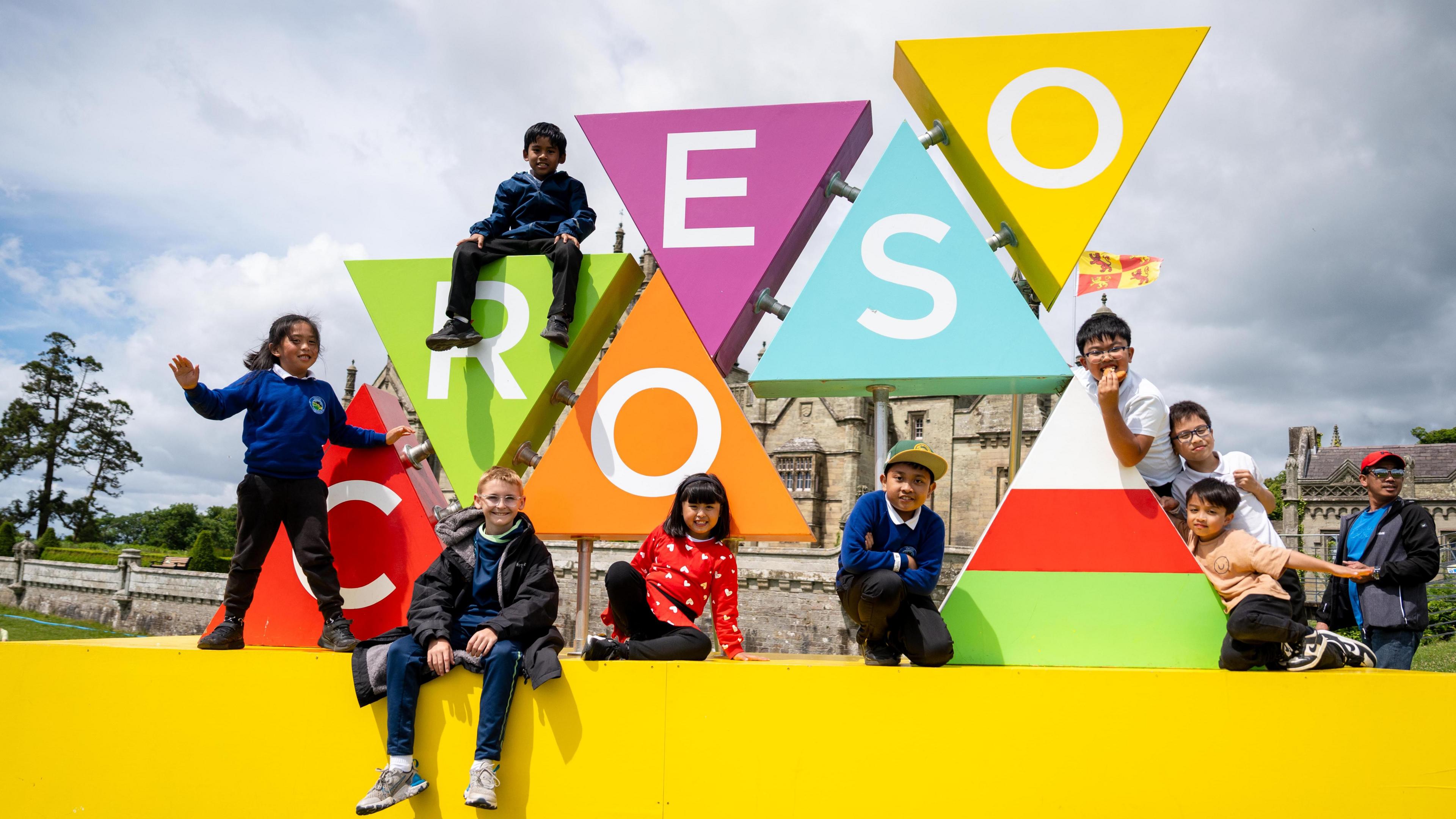 Wyth o blant, merched a bechgyn, yn eisted neu'n sefyll ar arwydd Croeso Eisteddfod yr Urdd