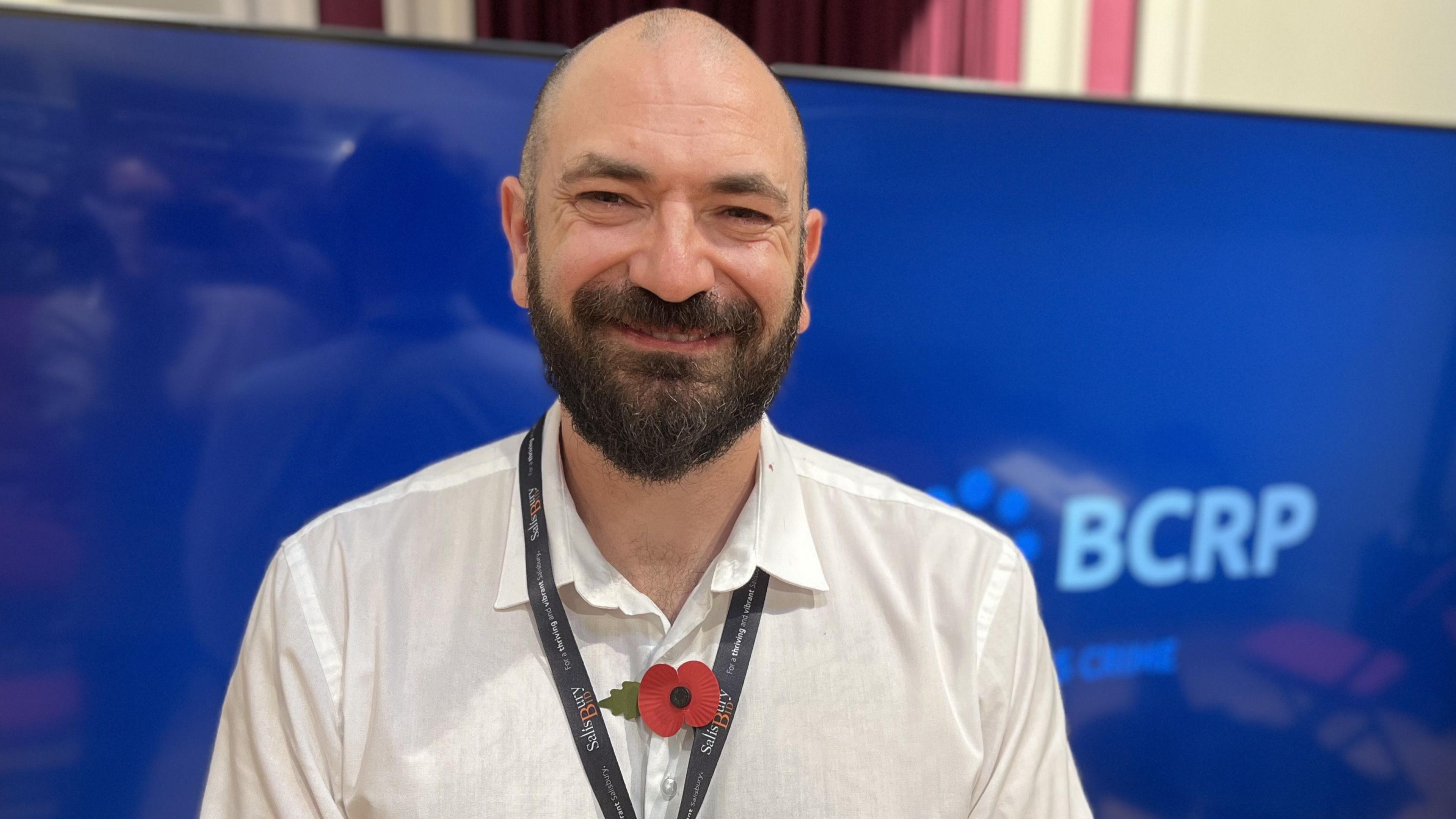 Dan Collins smiling with a brown and grey beard and a bald head, wearing a white shirt with a Remembrance poppy