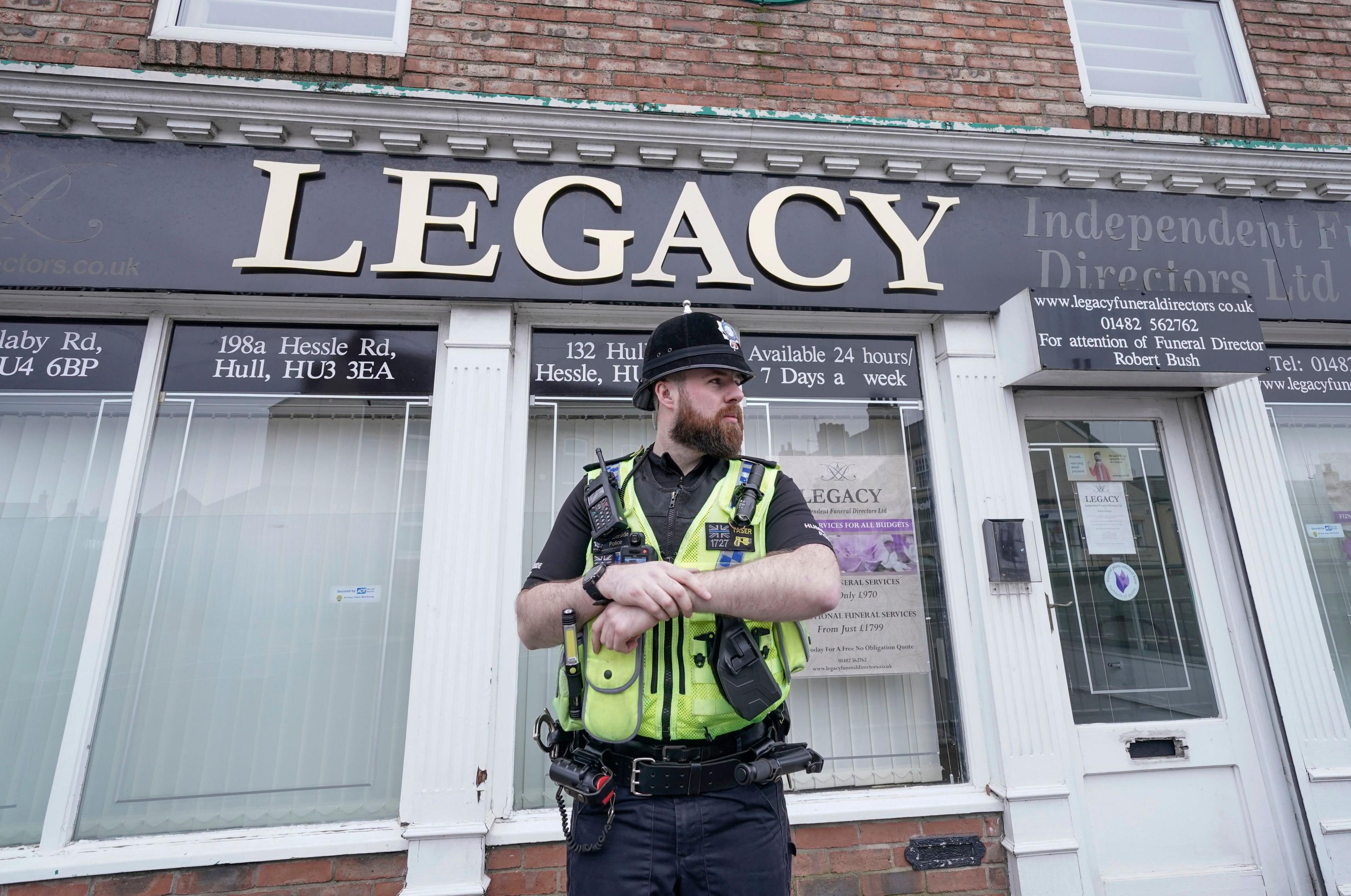 A police officer, wearing traditional uniform and a high-visibility vest, stands outside the Legacy Independent Funeral Directors. The building has large glass windows with white frames and a black and gold sign.