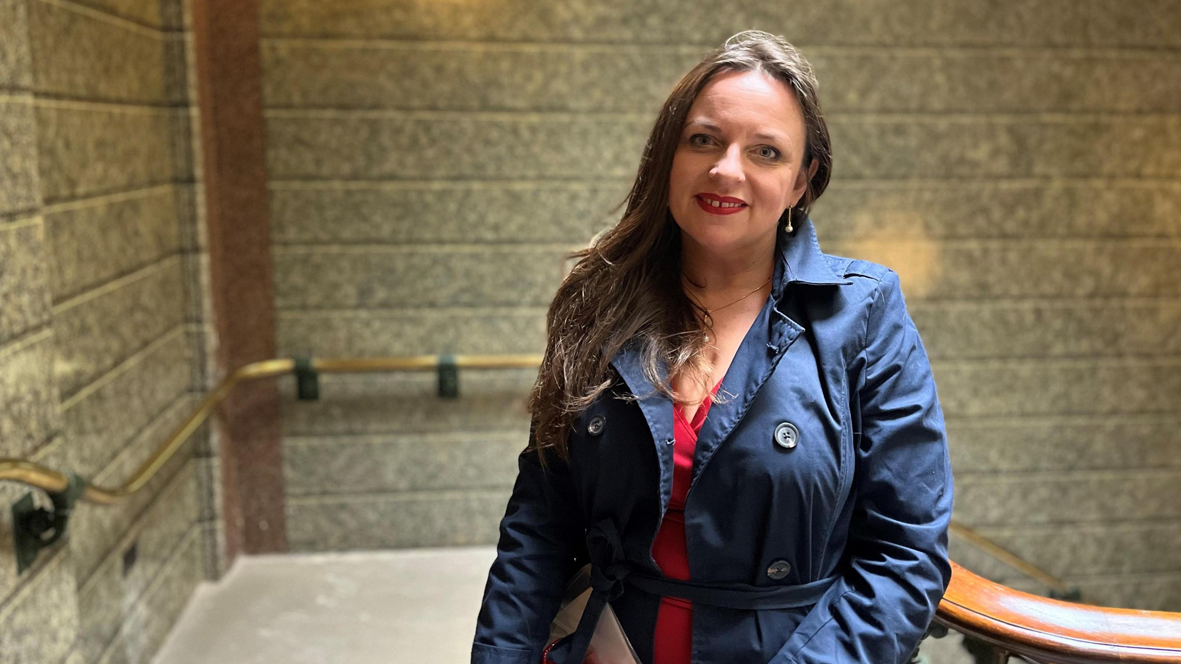 Angelika Kwidzinska is smiling as she stands on a sweeping staircase inside St George's Hall. She has long brown hair and is wearing a red dress with a navy blue raincoat.
