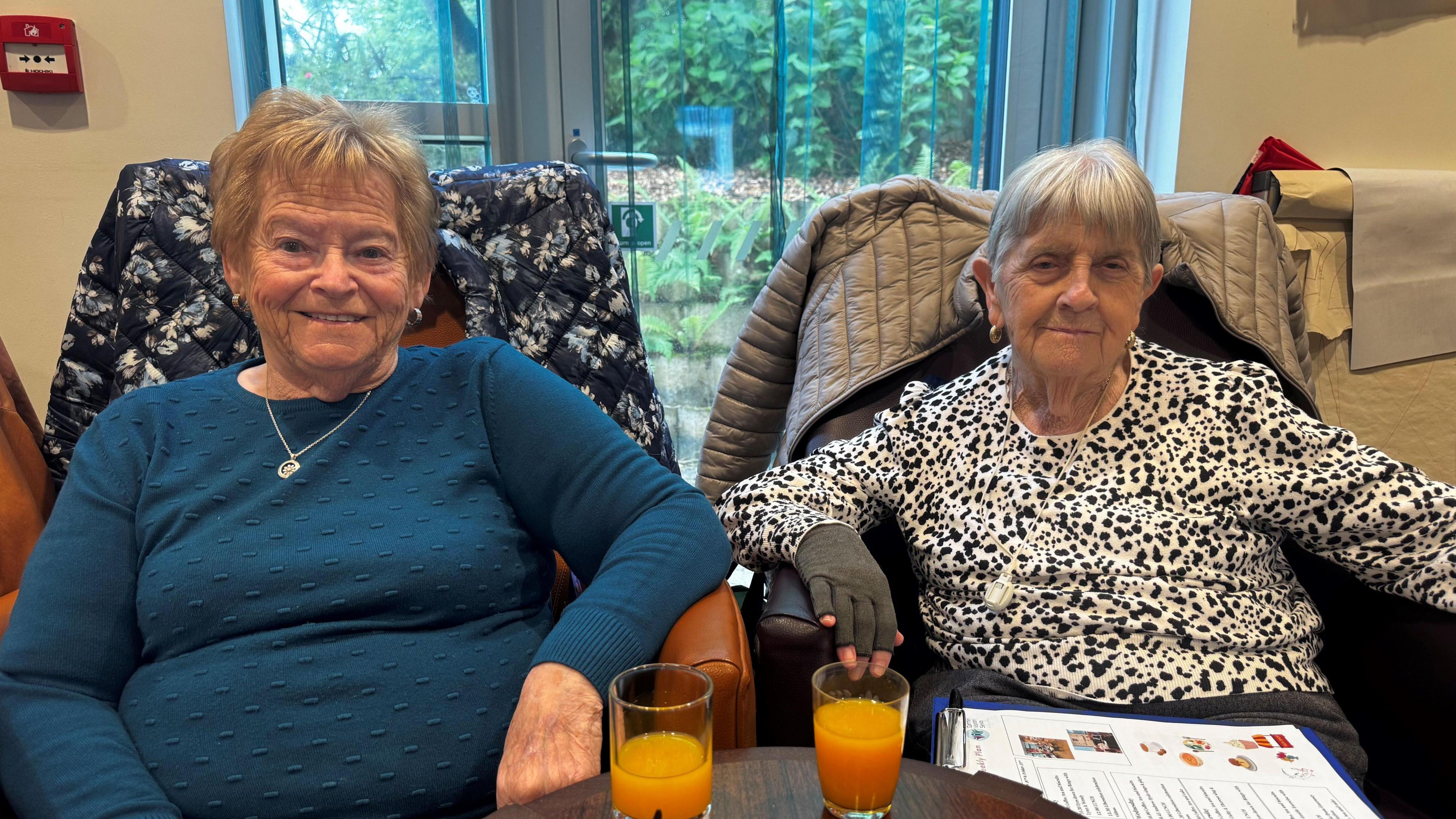 A photo of a lady with short, blonde hair and blue jumper sat in a chair next to a lady with short grey hair and a black and white speckled jumper.