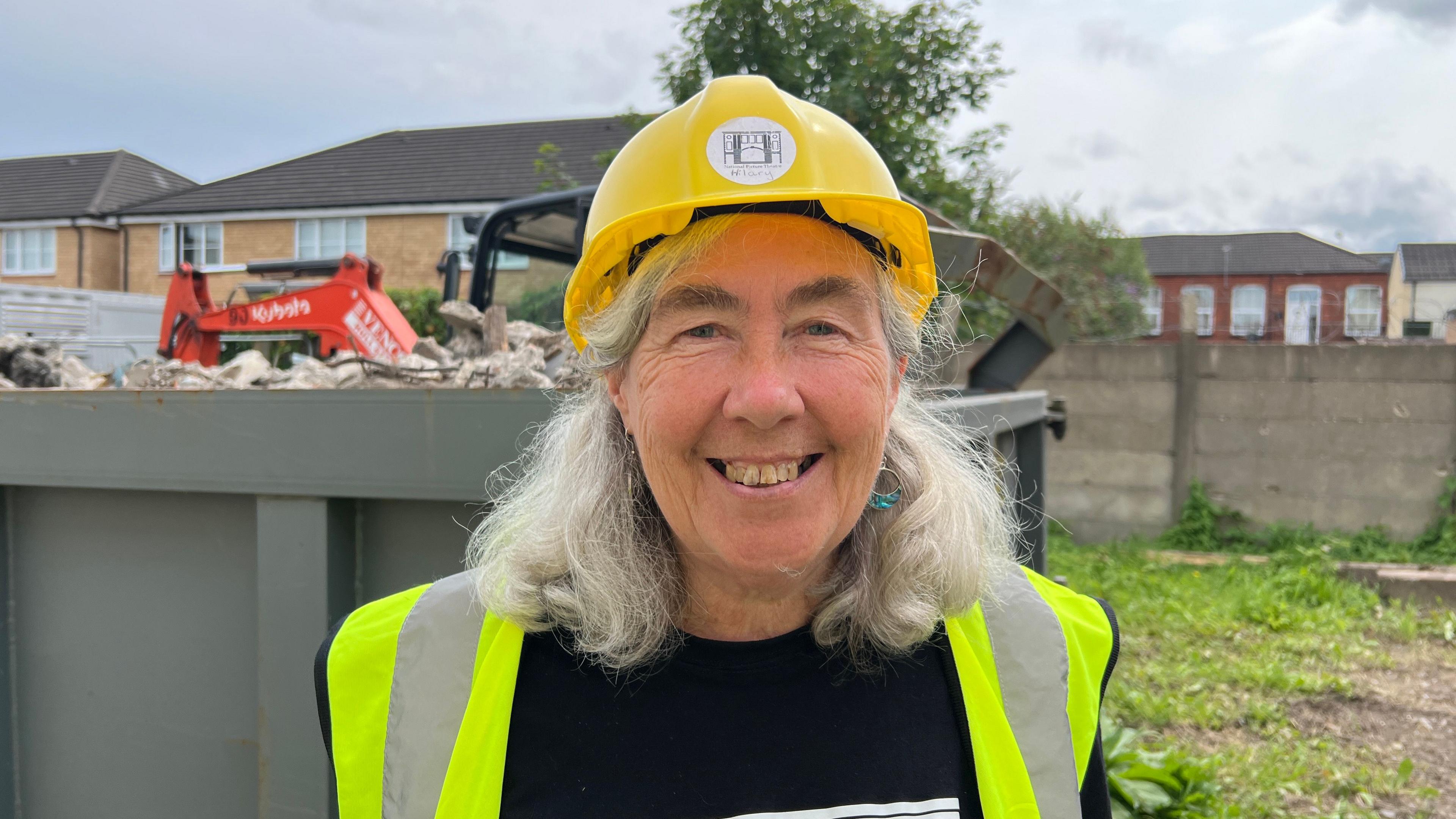 Hilary Byers stands on site at the theatre. She is wearing a yellow hard hat and a high-vis jacket. Rubble and construction machinery are in the background.