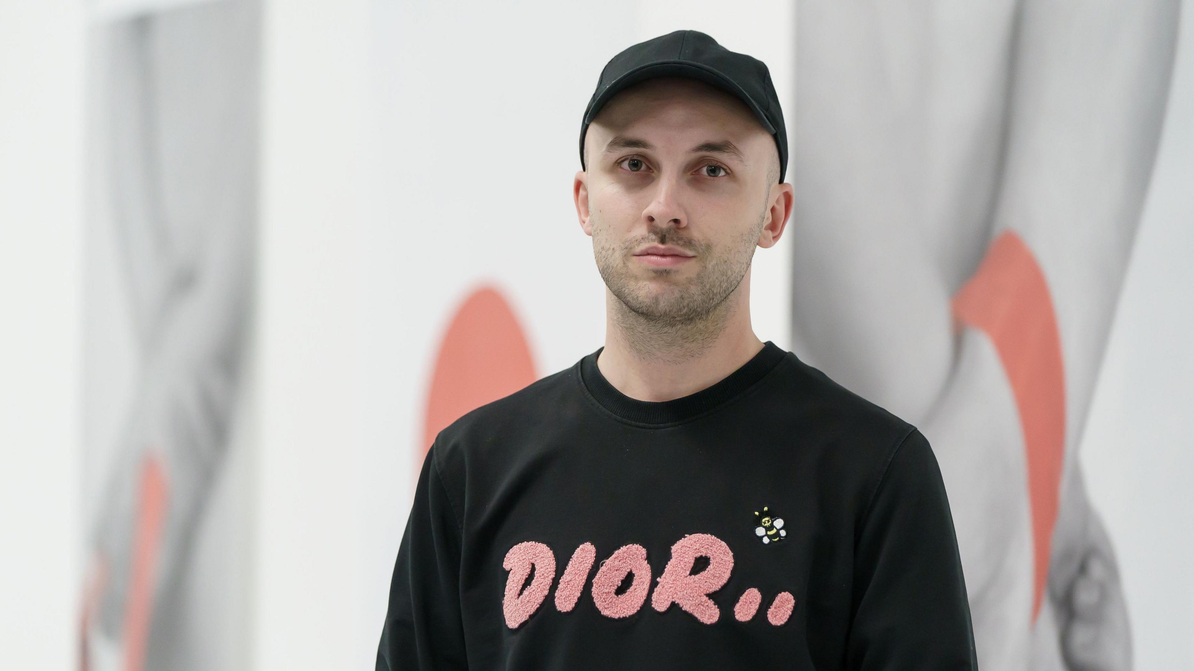 Dean Gooch looking into the camera with a straight face. He is wearing a black cap and a black jumper which has 'Dior..' written on it in pink letters. There is a bee above the lettering. He is standing in front of some artwork on a white wall which depicts hands.