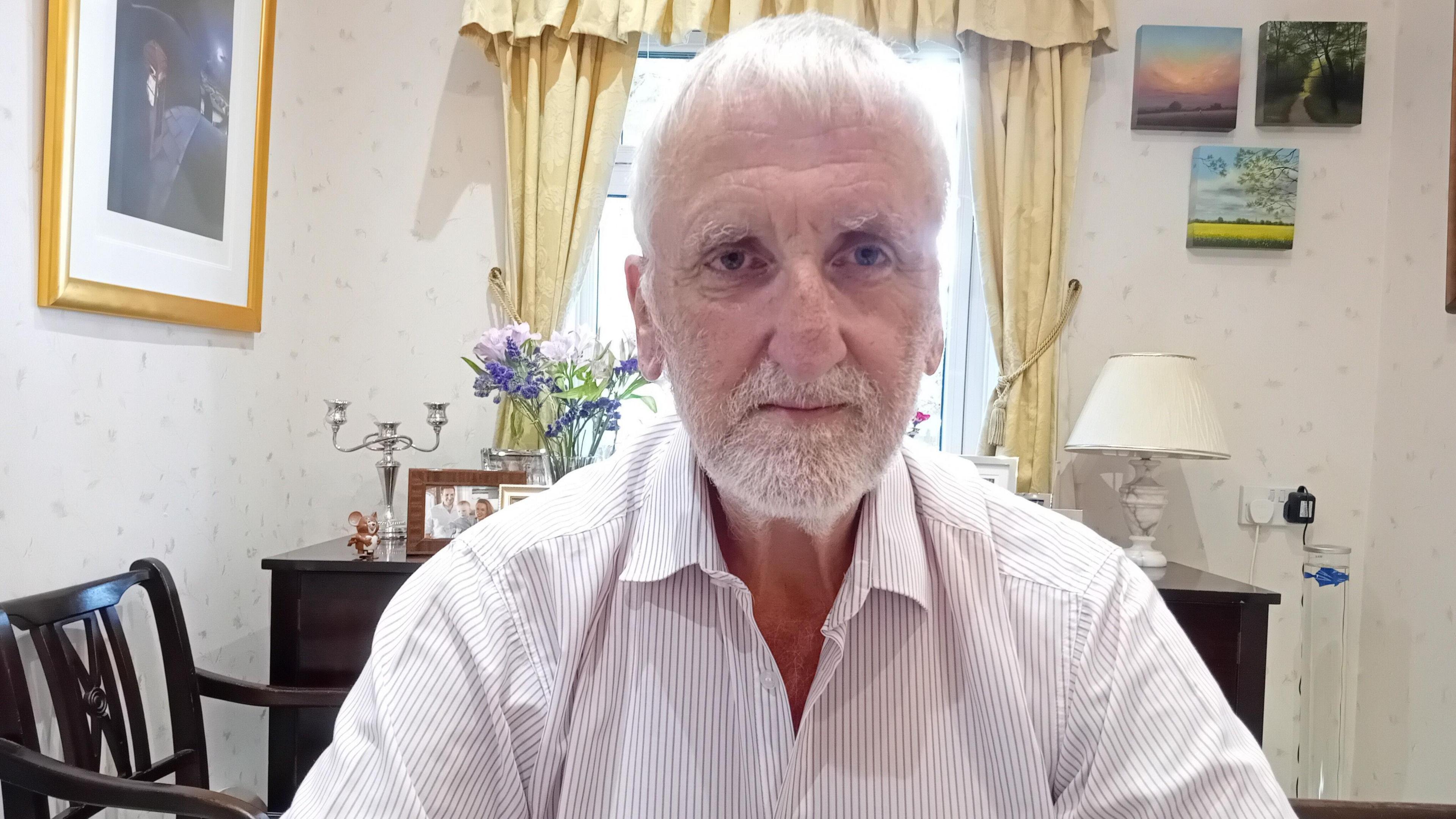 A man with white hair and white facial hair. He is wearing a white shirt with blue stripes. Behind him is a window with yellow curtains, and several pictures hung on the wall