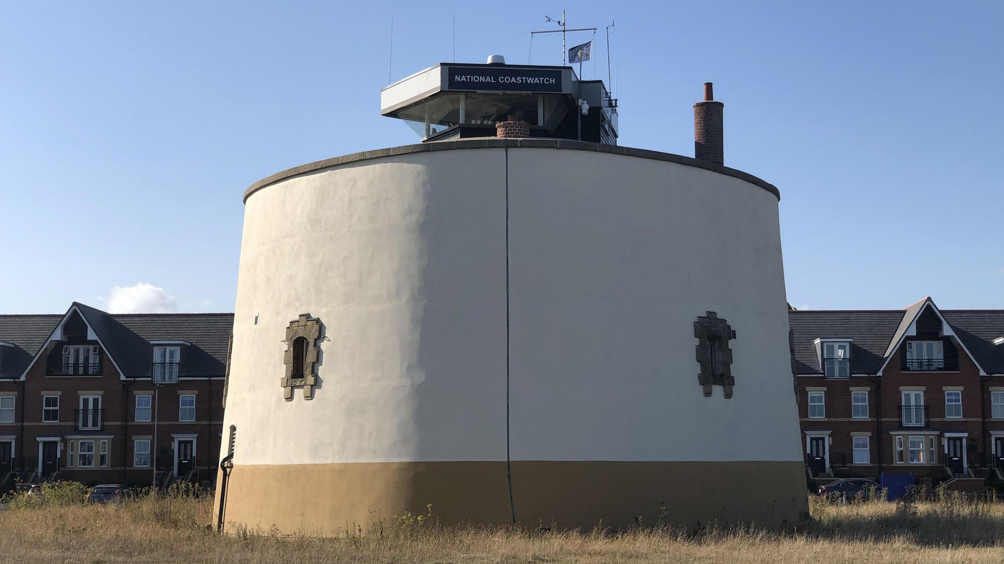 The round grey exterior of Martello Tower P in Felixstowe featuring small windows reinforced with bricks and a National Coastwatch lookout on top 