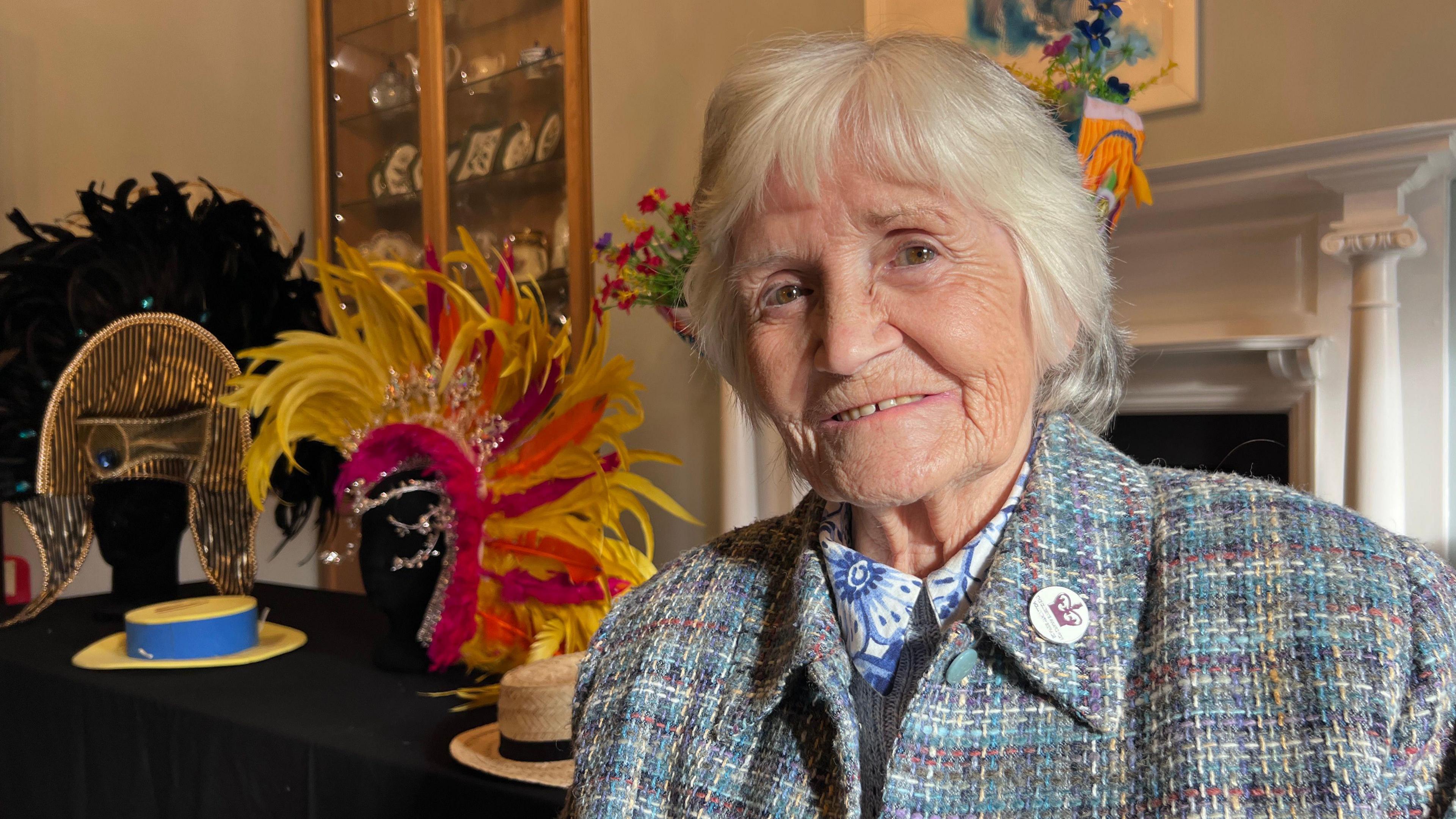 A close up of Marilyn Gearing, who had a stall at the first Luton Carnival in 1976. She is sat down in a room with colourful head pieces and hats behind her from previous carnivals. She is smiling and is wearing a coat featuring a badge with a crown on it.