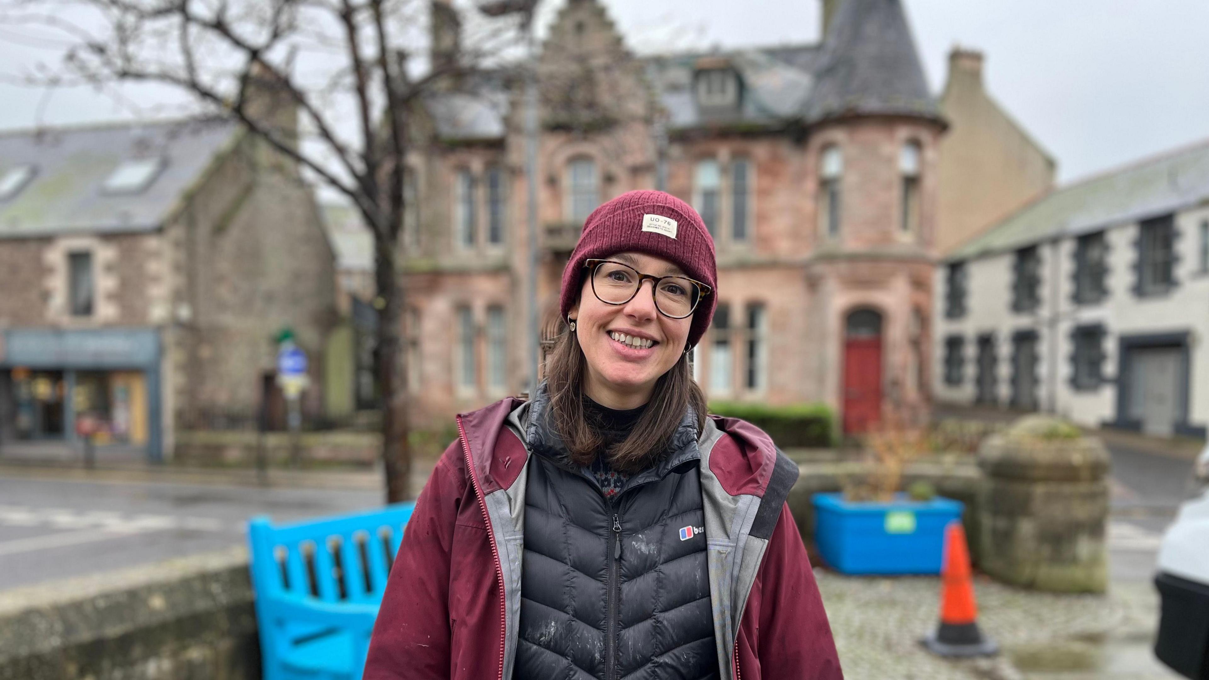 A woman with glases smiles at the camera. She's wearing a maroon bobble hat, black top and wine-coloured waterproof