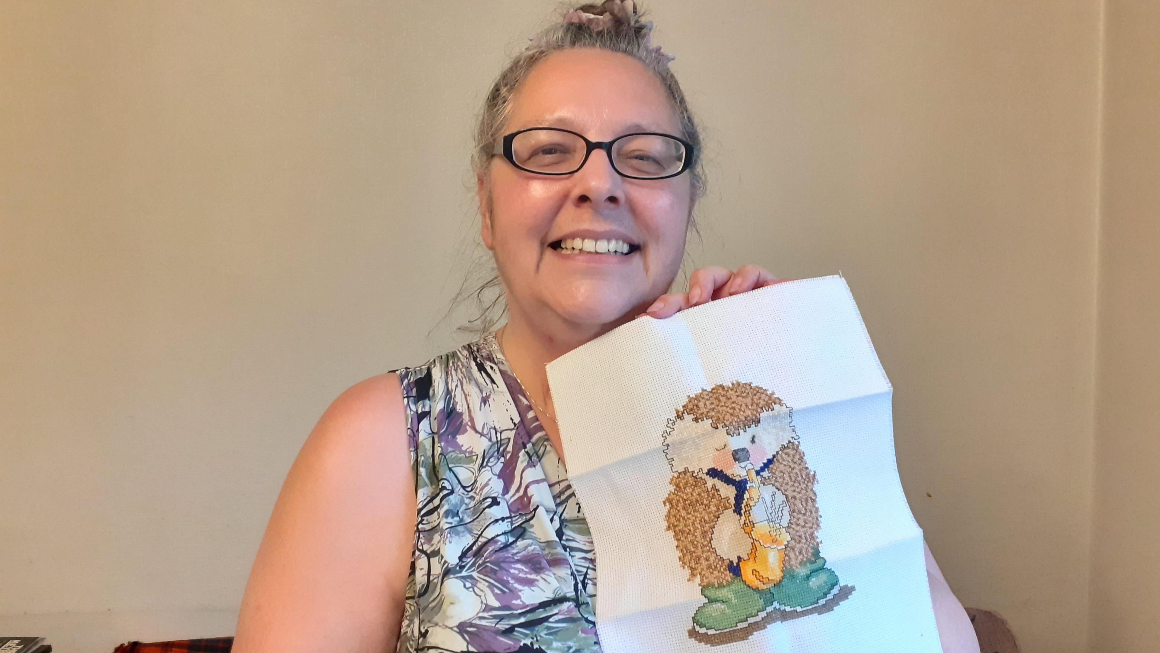 Molly Lofas, who has glasses and tied-up grey hair, smiles as she holds up a cross-stitch of a hedgehog in a blue tie and green wellington boots playing a saxophone