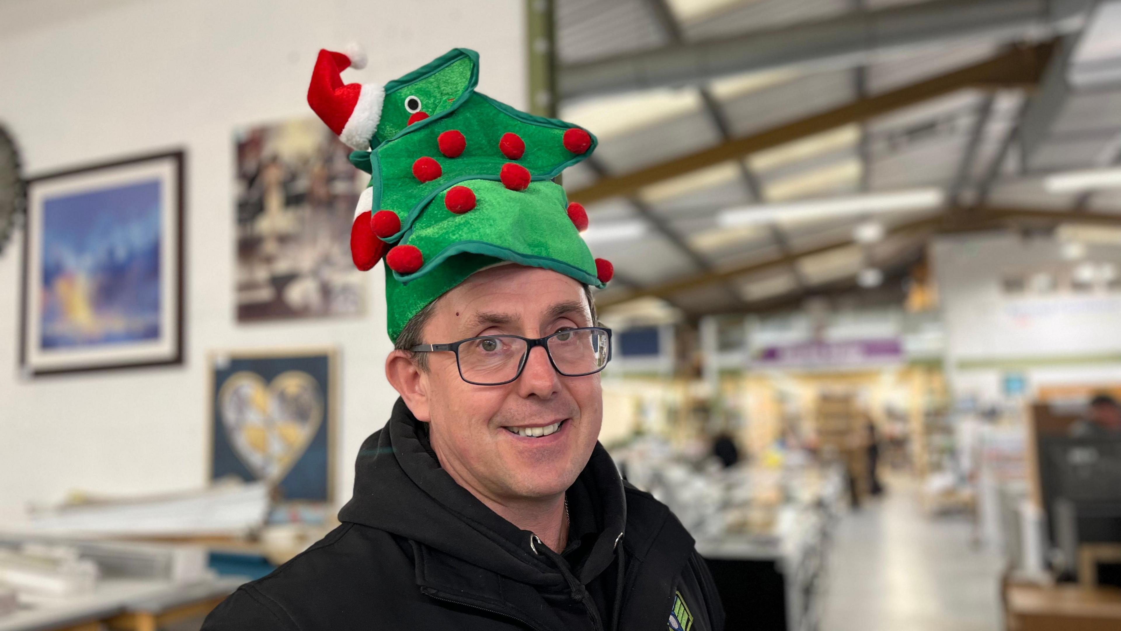 Daniel Thompson smiles at the camera. He is wearing glasses and a black hoodie. He is wearing a novelty Christmas tree hat. Blurred behind him is a warehouse full of furniture and appliances.