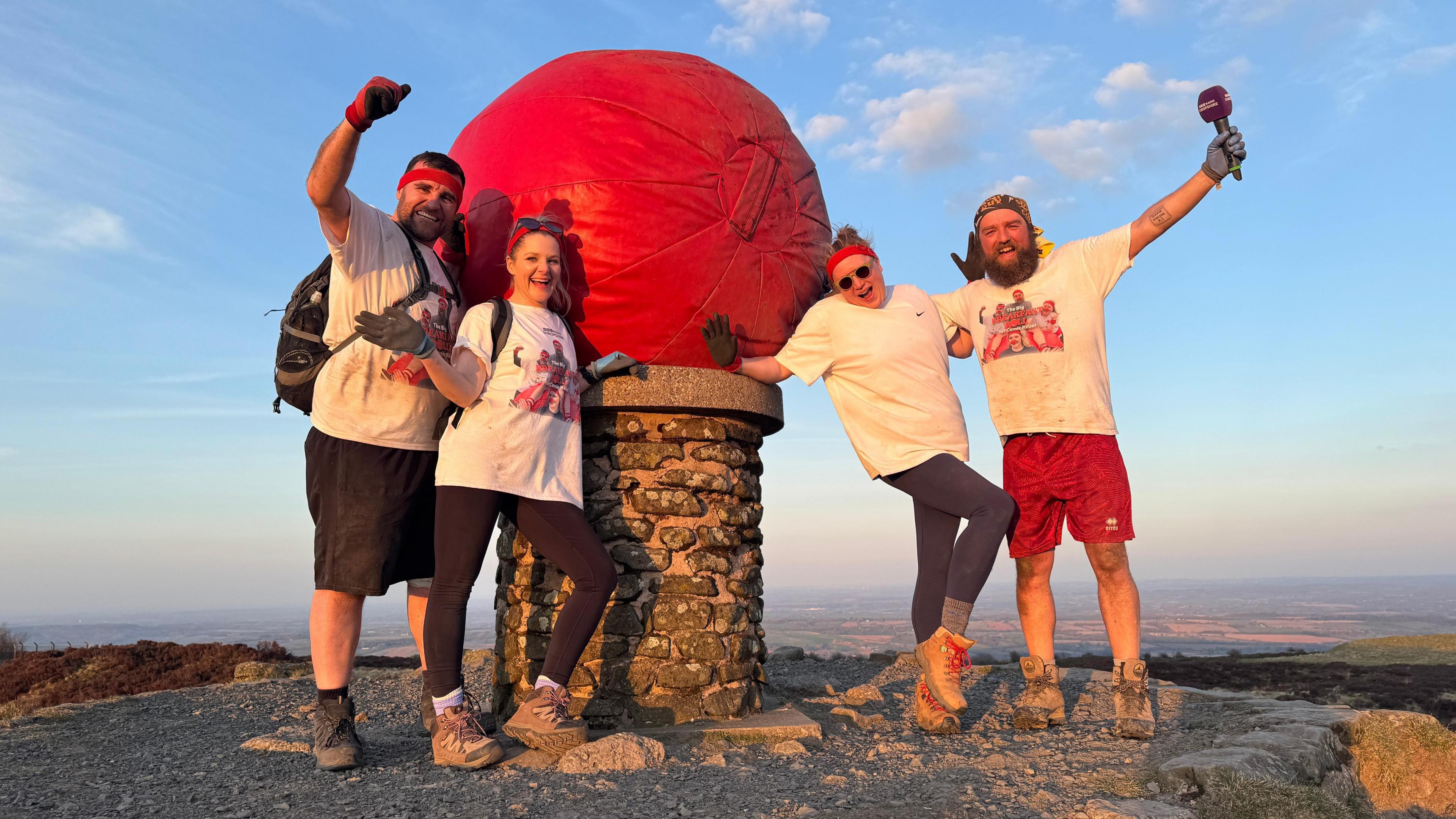 Four people stand on top of a large hill that looks out across an expanse of land. There is a huge red ball on a podium between them.