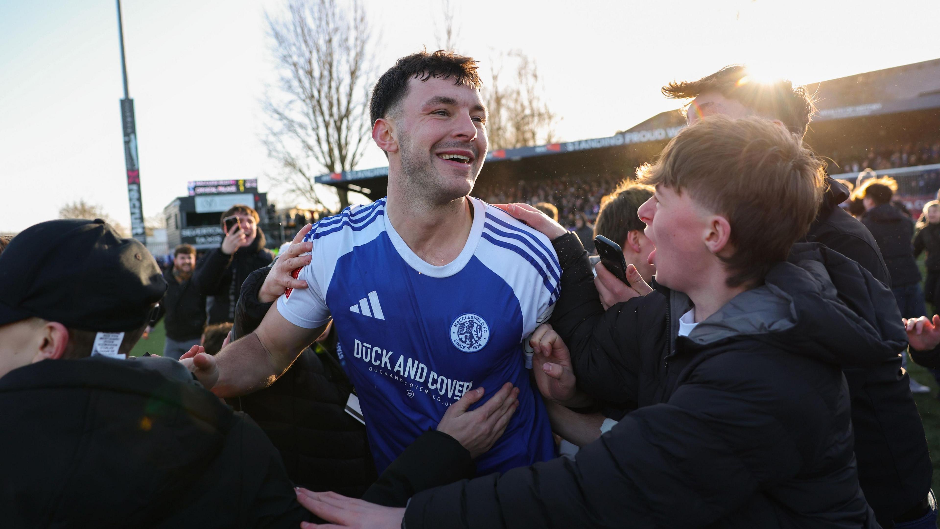 A smiling Sam Heathcote, in blue and white Macclesfield shirt, is surrounded by excited fans during a pitch invasion