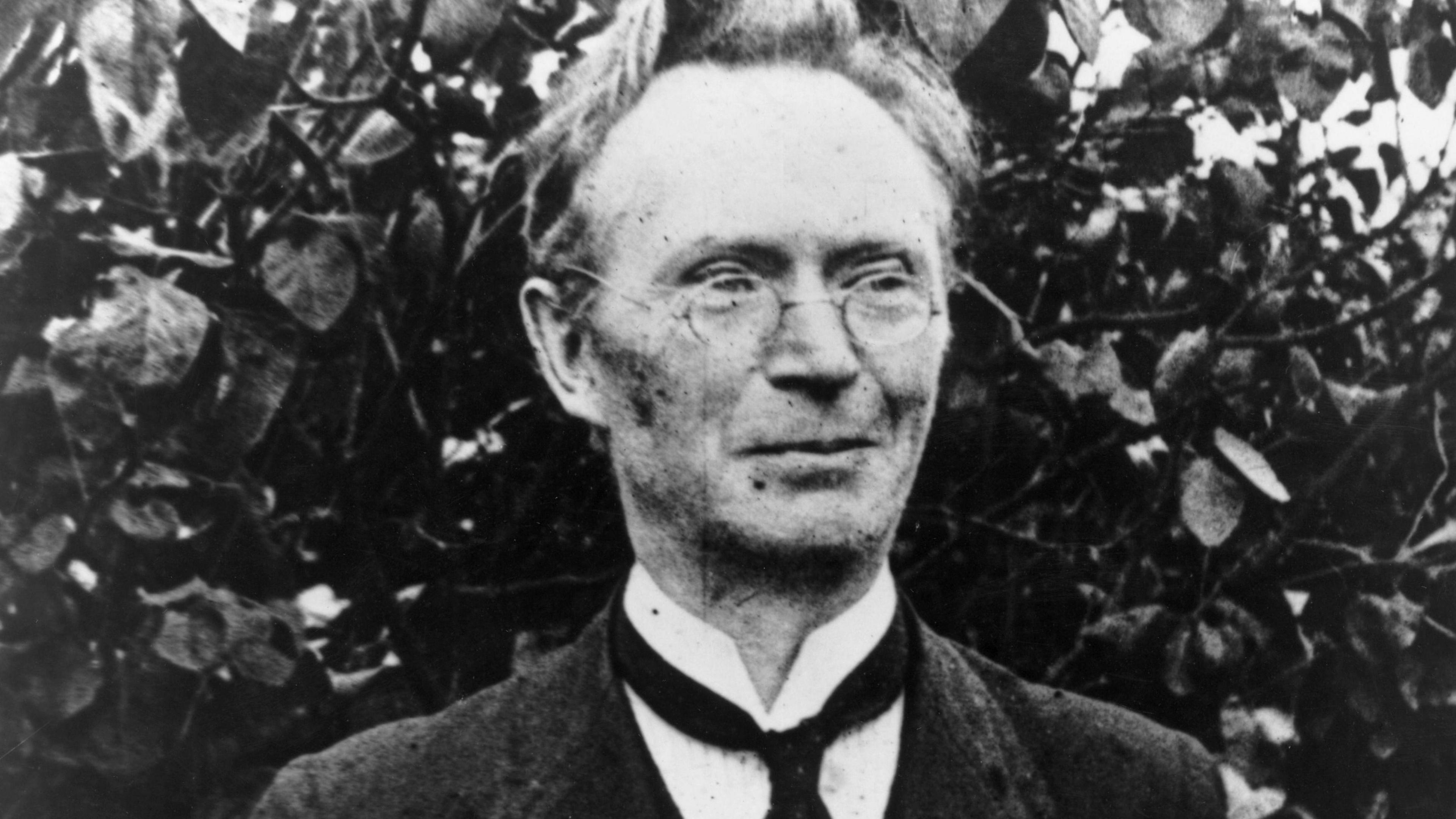 A black and white archive photo of Eoin MacNeill circa 1910.  He had short, receeding, fair hair and small round-rimmed glasses.  He was wearing a dark suit, a white shirt and black cravat-style tie.