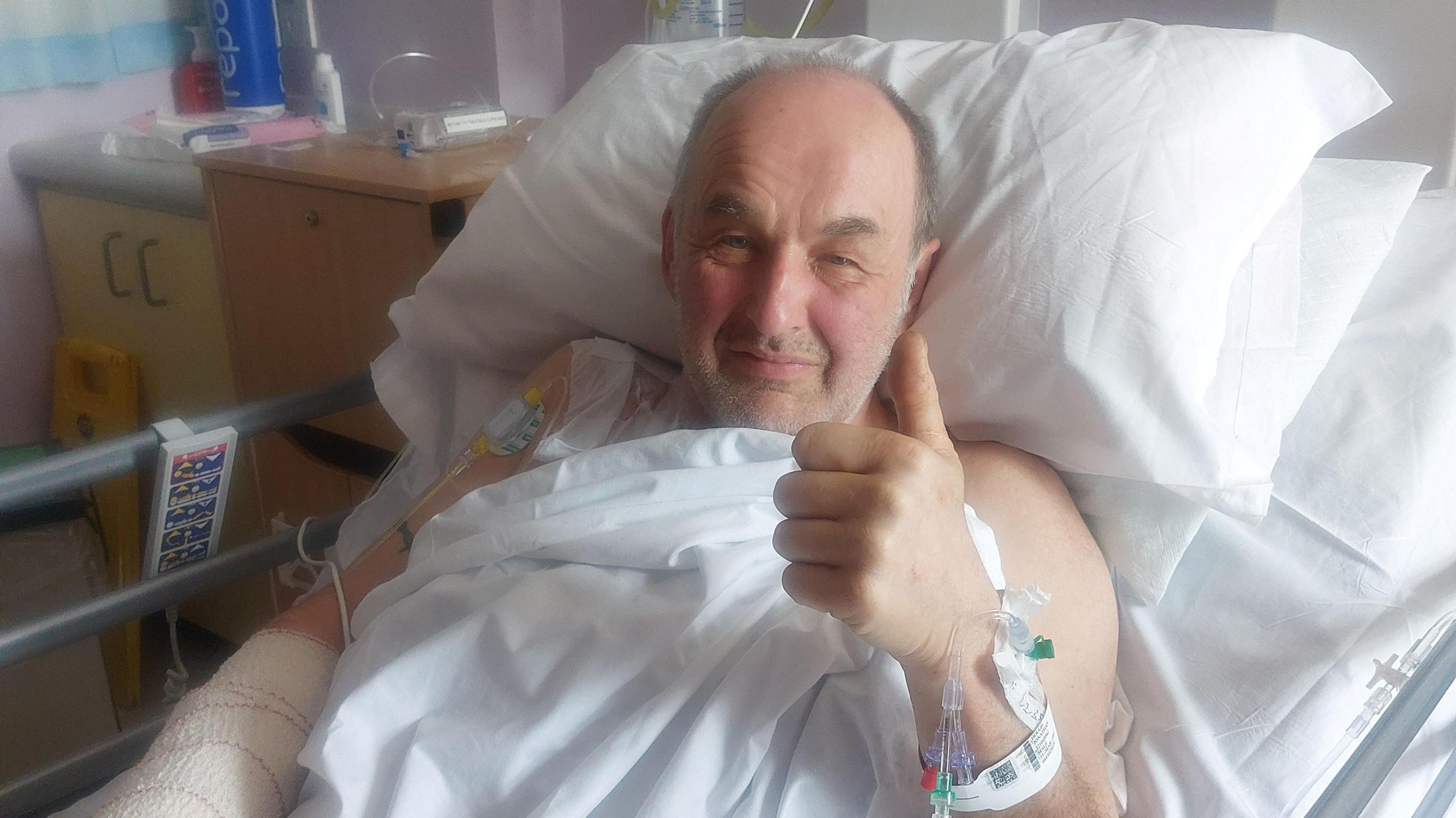 Joe Hughes, lying in a hospital bed, holding up his left hand, with medical equipment in his arms, sitting on white sheets and pillows, and smiling at the camera. He has short grey hair and a stubbly beard. There are two hospital cabinets to the right.