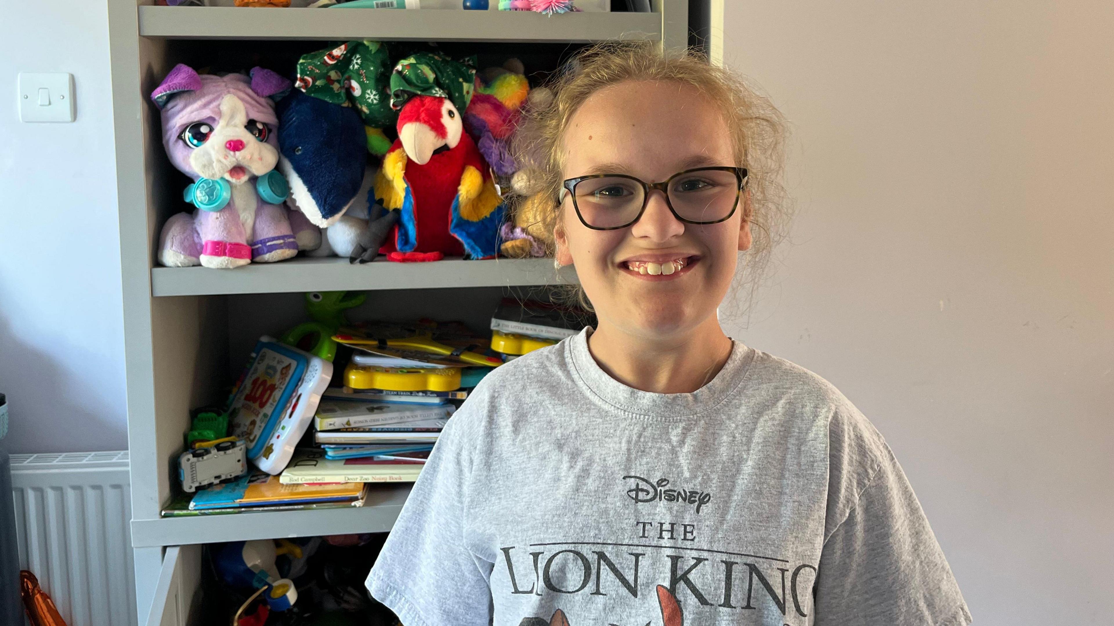 A girl with blonde hair and glasses wearing a grey Lion King T-shirt. She is standing in front of a book case filled with stuffed toys and books.