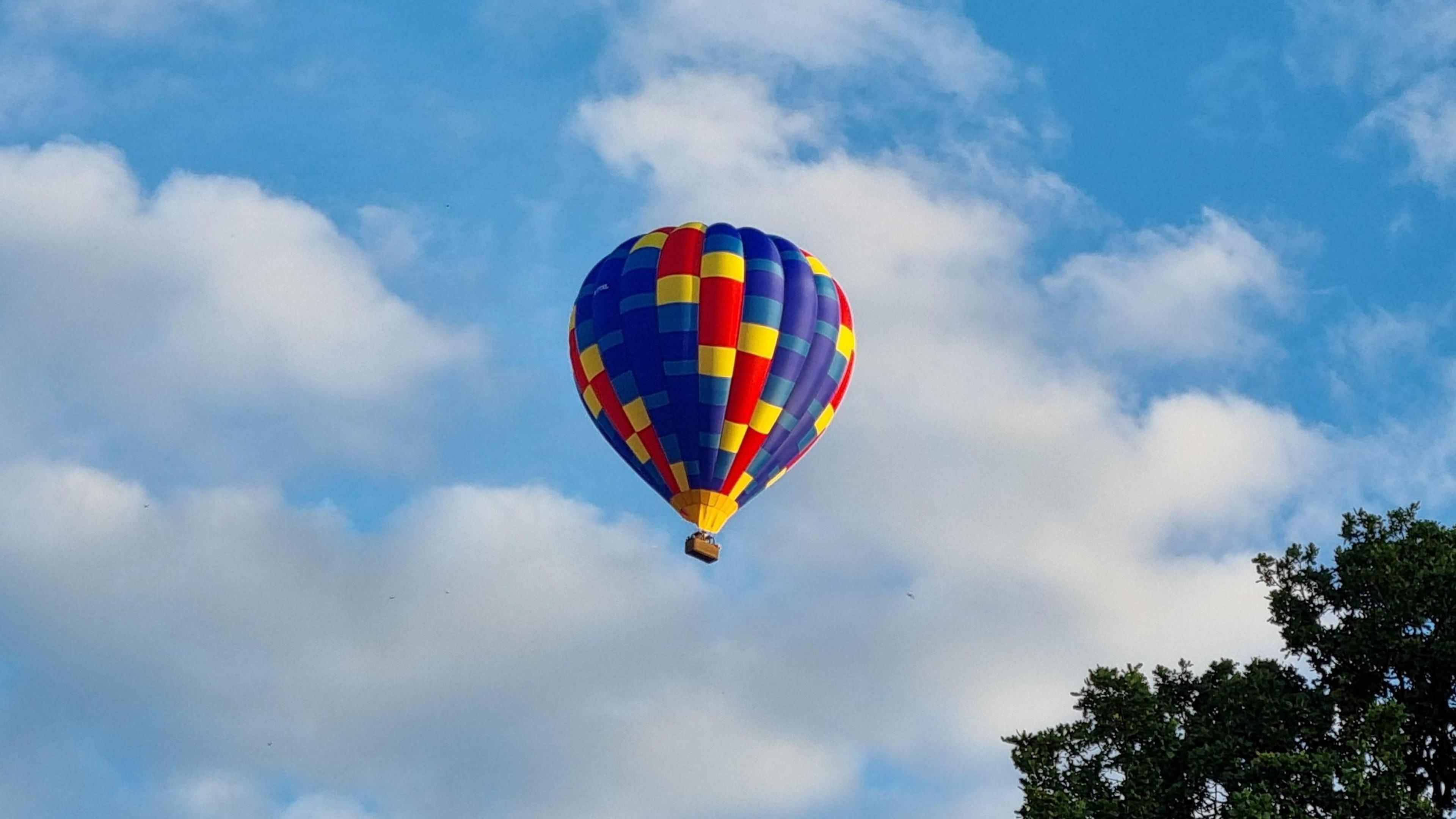 A hot-air balloon, coloured in irregular red, yellow and blue panels, floats in a blue sky with white clouds. The top of a tree is visible bottom-right. 