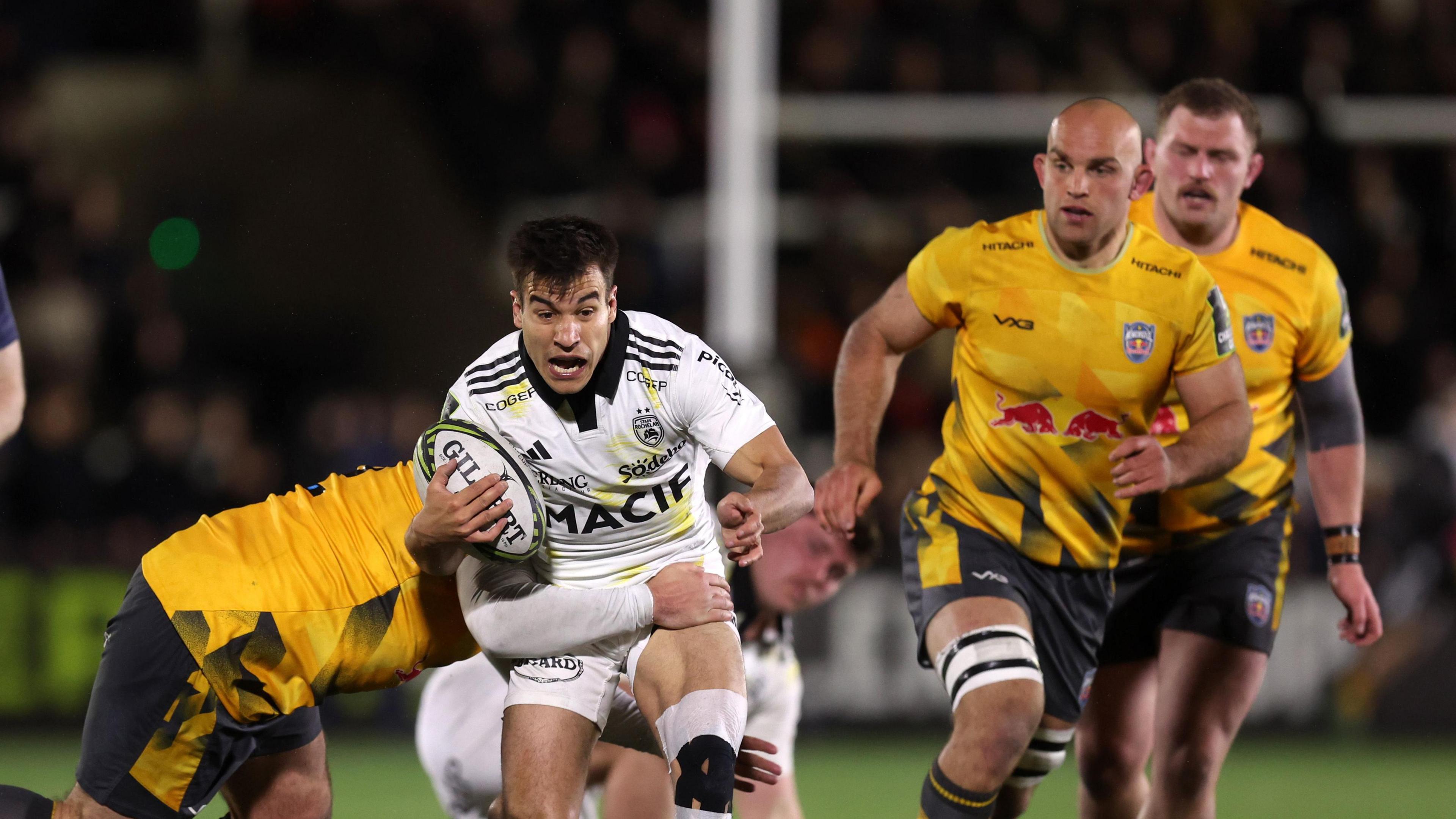 La Rochelle scrum-half Thomas Berjon carrying the ball but is tackled by a Newcastle Red Bulls player whose face is obscured, with two other Red Bulls players watching on the right