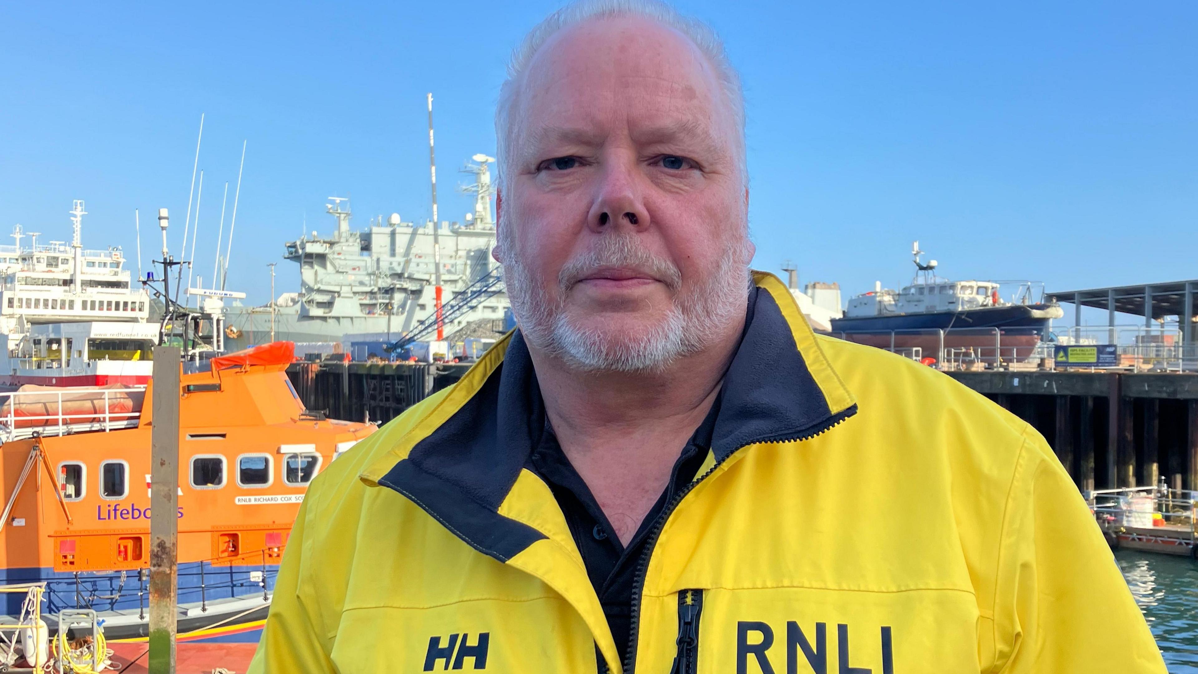 Deputy second coxswain David Nicholl stands in front of the retired lifeboat Richard Cox Scott. He is dressed in a yellow jacket with the RNLI logo on it. 