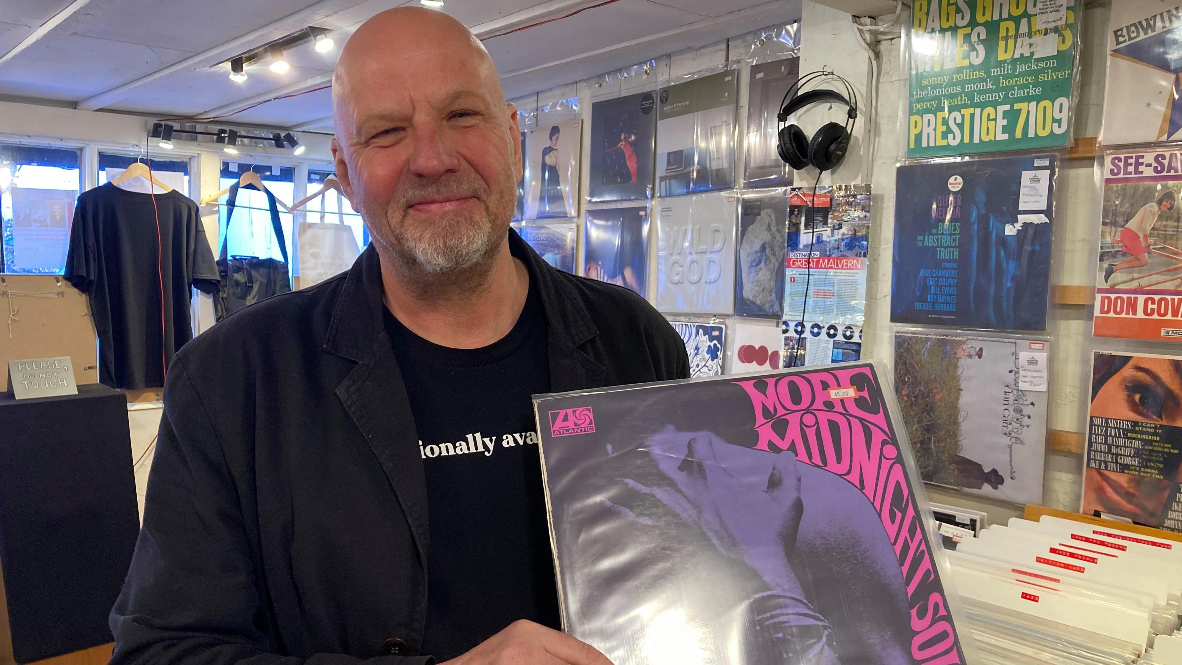 A man with a shaved head and a grey beard stands in a shop holding a vinyl record in its sleeve. There are other vinyl records mounted on the wall behind.