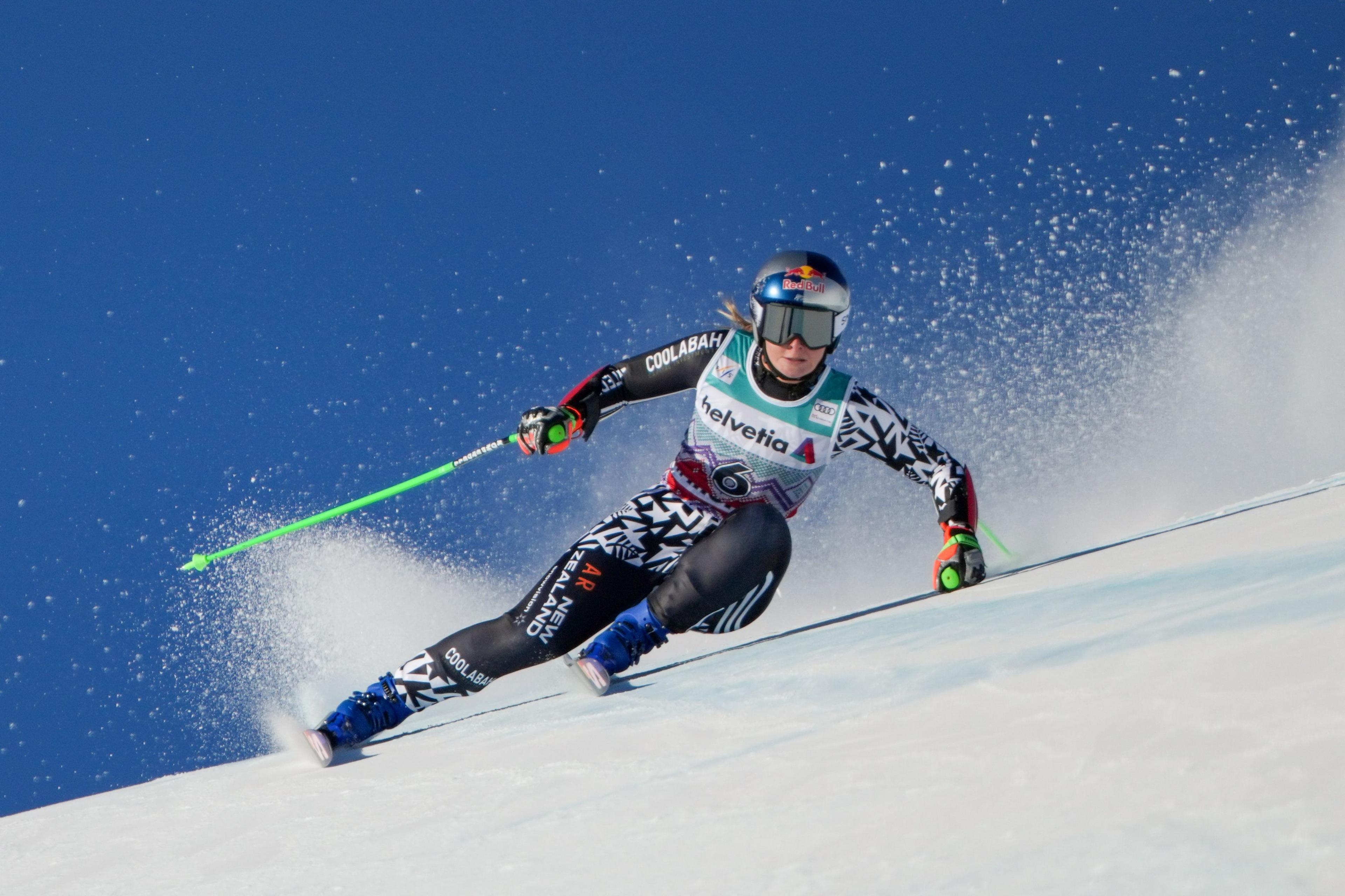 A downhill skier in a patterned racing suit leans aggressively into a high‑speed turn on a snowy slope, carving a sharp edge as a spray of snow trails behind under a clear blue sky.